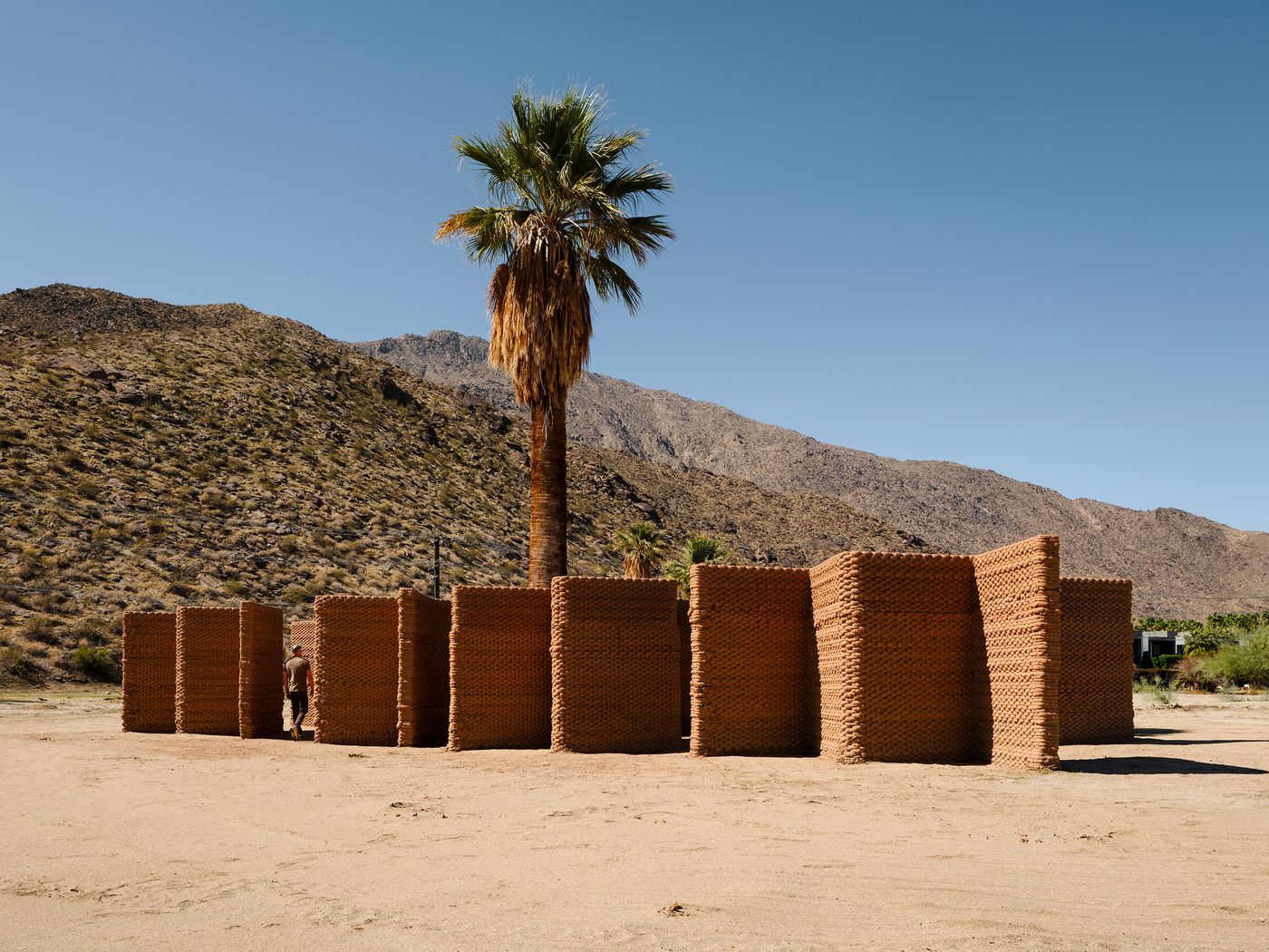 A person explores a structure made of stacked bricks near a palm tree, set against a desert landscape and mountainous backdrop. A person explores a structure made of stacked bricks near a palm tree, set against a desert landscape and mountainous backdrop.