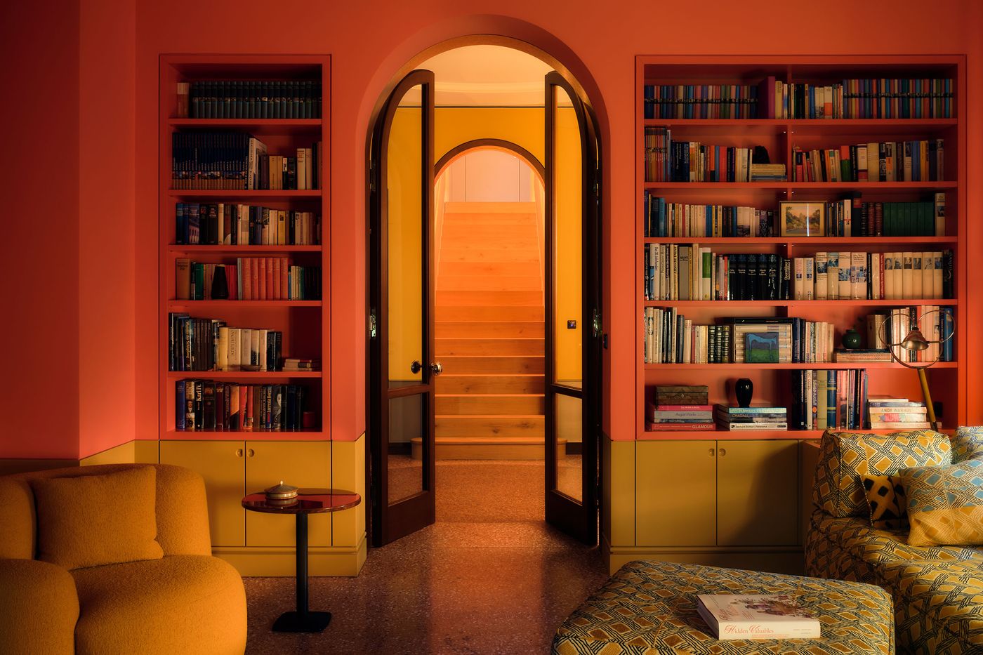 A cozy reading nook with vibrant pink walls, bookshelves, and a stylish yellow couch, leading to wooden stairs and a warm hallway. A cozy reading nook with vibrant pink walls, bookshelves, and a stylish yellow couch, leading to wooden stairs and a warm hallway.