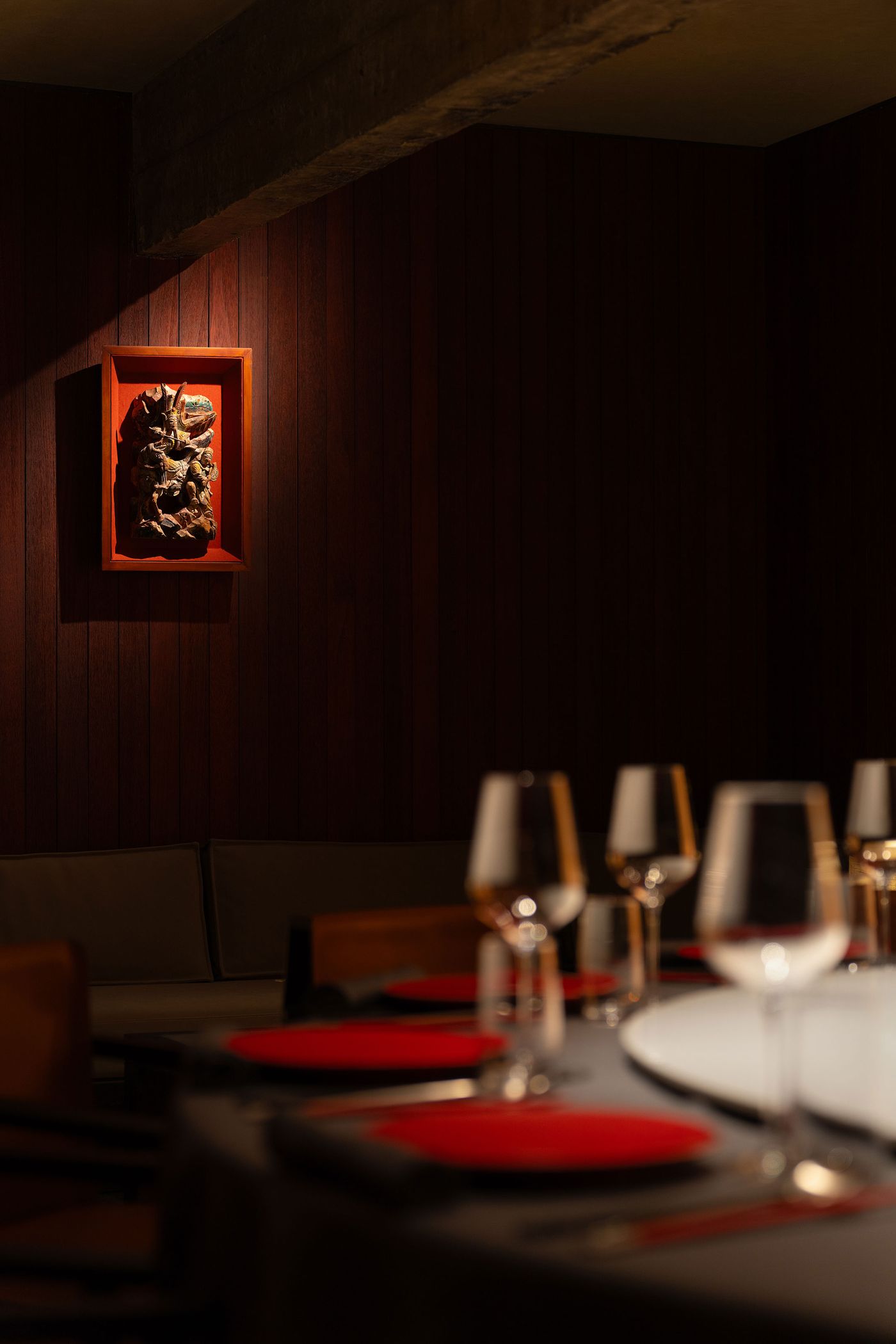 Dimly lit restaurant interior featuring a wooden wall, a carved artwork, and an elegantly set table with red placemats and glassware.