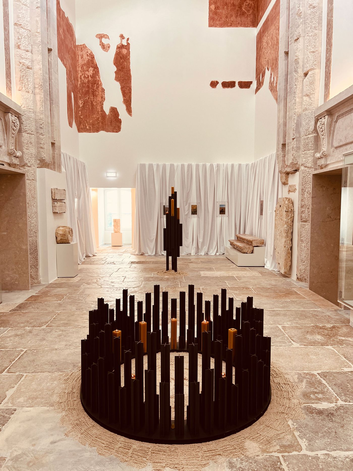 An atmospheric courtyard at Locke Santa Joana, featuring a sculptural installation by AB and AC Architects. Titled "Alma Mater", is consists of two large-scale pieces, composed of dark, vertical elements that stand out amid the historic stone-floored space. Stripped walls with faded murals and white drapes hint at preserved architectural history, creating a meditative blend of old and new. An atmospheric courtyard at Locke Santa Joana, featuring a sculptural installation by AB and AC Architects. Titled "Alma Mater", is consists of two large-scale pieces, composed of dark, vertical elements that stand out amid the historic stone-floored space. Stripped walls with faded murals and white drapes hint at preserved architectural history, creating a meditative blend of old and new.