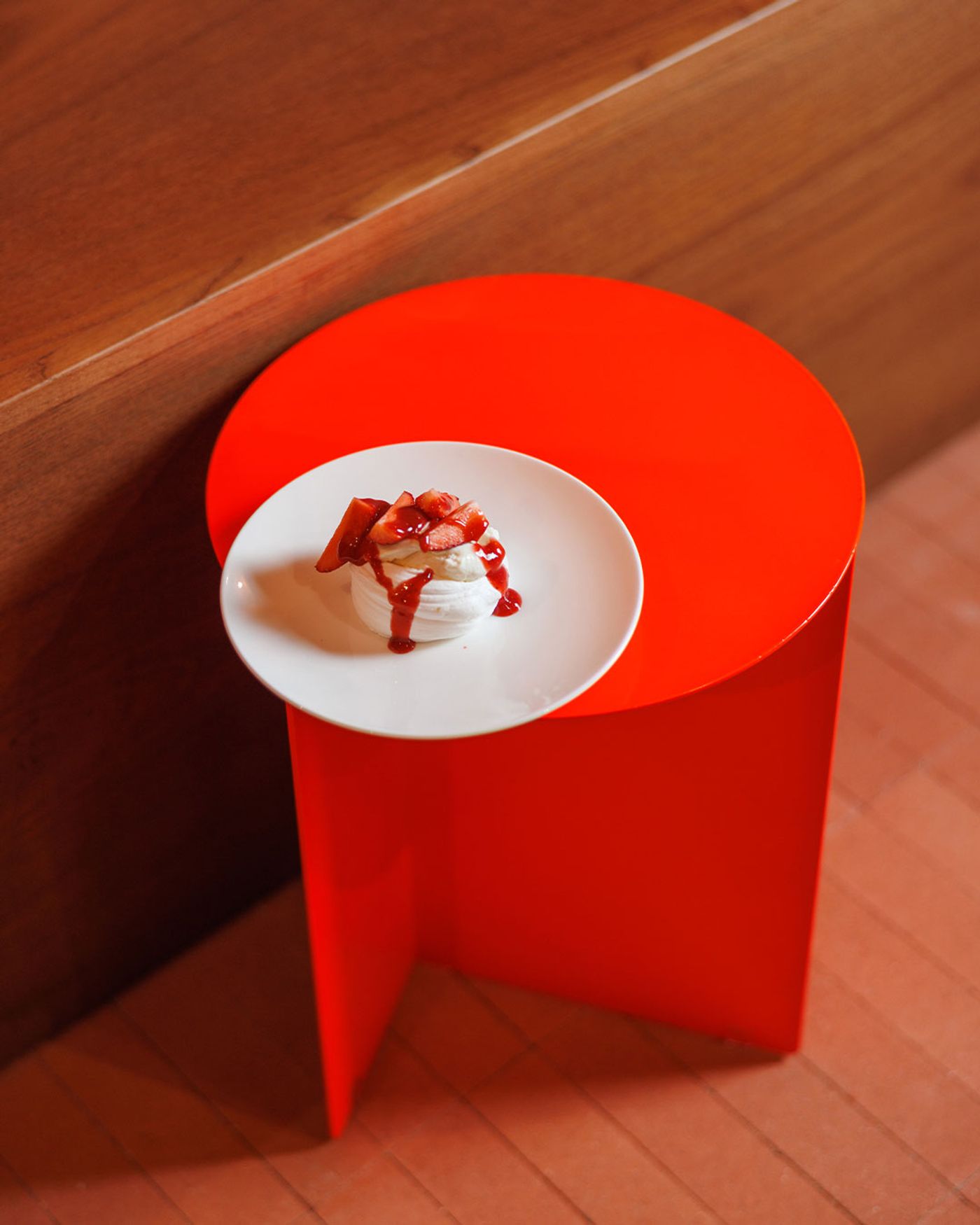 A beautifully plated dessert from Santo Palato, featuring a pristine white meringue topped with fresh strawberries and a vibrant red coulis, artfully placed on a small, striking red cylindrical table. The composition, set against a warm wooden backdrop, highlights the dessert's freshness and the restaurant's playful use of color.