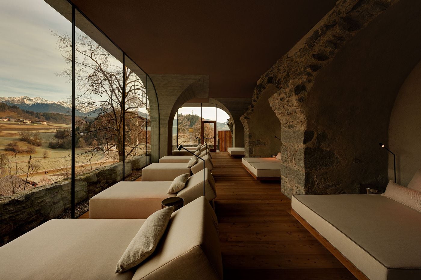 A spa relaxation room at Castel Badia sets cream loungers along a full-height glass wall above a medieval stone parapet, facing snow-capped peaks. Opposite, rough rubble masonry and arched openings meet board-marked concrete arches and timber floors, a measured dialogue between old fabric and new intervention. A spa relaxation room at Castel Badia sets cream loungers along a full-height glass wall above a medieval stone parapet, facing snow-capped peaks. Opposite, rough rubble masonry and arched openings meet board-marked concrete arches and timber floors, a measured dialogue between old fabric and new intervention.