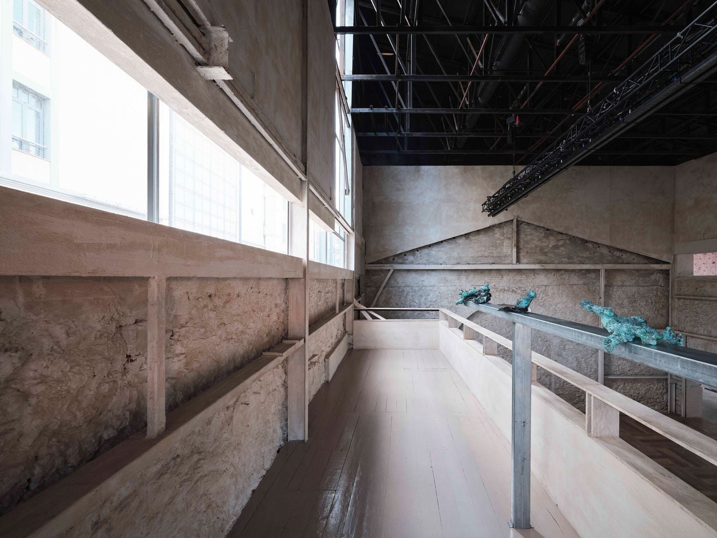 An elevated walkway runs alongside the retained masonry wall, its pale timber floor and restrained balustrade contrasting with the rough historic fabric. Sculptural works displayed along a slim metal rail animate the circulation route, while the exposed black roof structure reinforces the gallery’s industrial character. An elevated walkway runs alongside the retained masonry wall, its pale timber floor and restrained balustrade contrasting with the rough historic fabric. Sculptural works displayed along a slim metal rail animate the circulation route, while the exposed black roof structure reinforces the gallery’s industrial character.