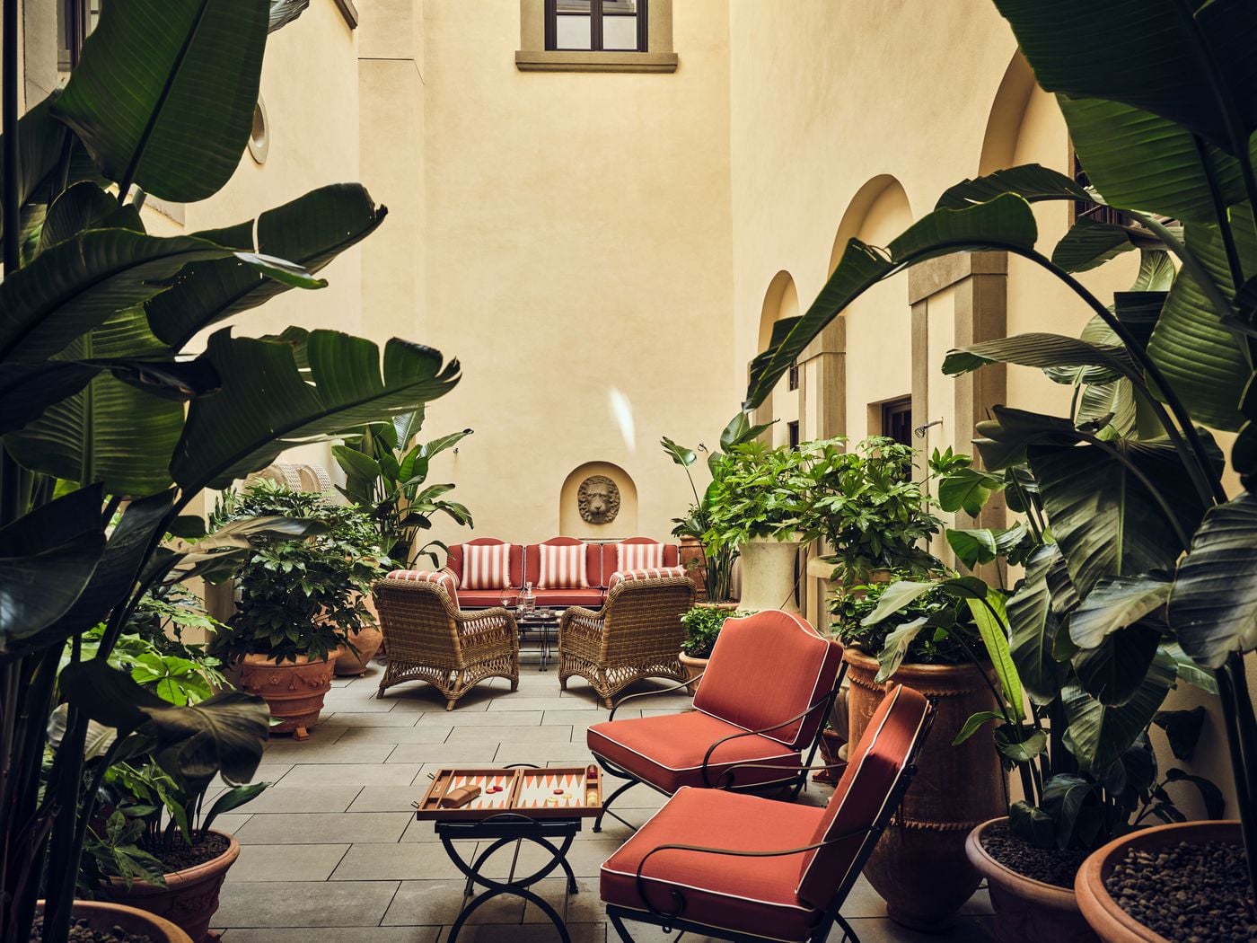 A wider view of the courtyard at La Réserve Firenze, framed by the broad leaves of potted banana plants. Coral cushioned iron chairs and a backgammon set occupy the foreground, while wicker armchairs and a striped sofa gather beneath the lion's-head fountain, enclosed by the palazzo's tall ochre walls and arched openings. A wider view of the courtyard at La Réserve Firenze, framed by the broad leaves of potted banana plants. Coral cushioned iron chairs and a backgammon set occupy the foreground, while wicker armchairs and a striped sofa gather beneath the lion's-head fountain, enclosed by the palazzo's tall ochre walls and arched openings.