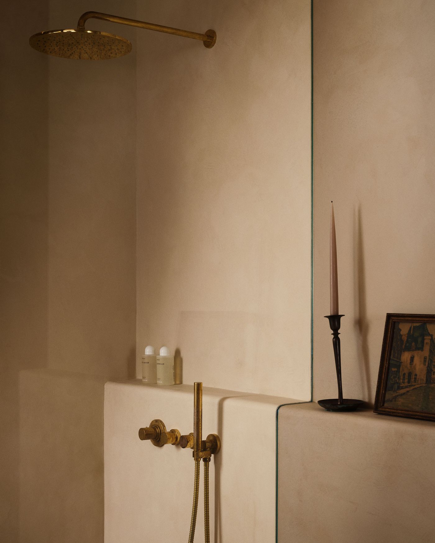 A luxurious shower space featuring a gold showerhead, minimalist toiletries, and a candle beside an art piece on a sleek, neutral wall.