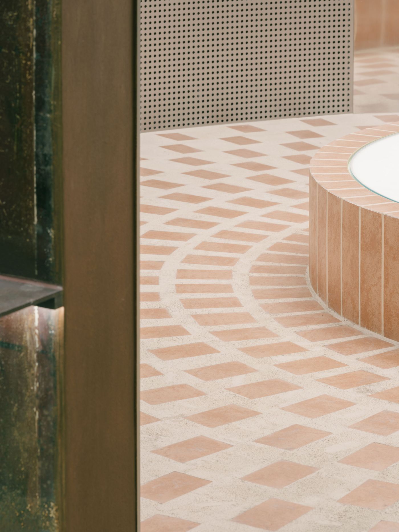 A striking circular pattern of terracotta tiles on a light concrete floor creates a visually dynamic pathway within AITASHOP, leading towards a perforated grey wall. In the foreground, a curved display structure clad in terracotta tiles with a white circular top suggests a dedicated presentation area, blending natural textures with modern lines. A striking circular pattern of terracotta tiles on a light concrete floor creates a visually dynamic pathway within AITASHOP, leading towards a perforated grey wall. In the foreground, a curved display structure clad in terracotta tiles with a white circular top suggests a dedicated presentation area, blending natural textures with modern lines.