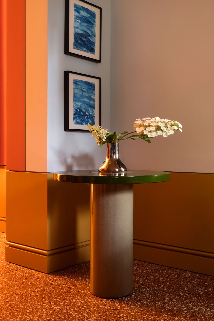 A modern table with a green top and metallic base, holding a vase of white flowers, near blue abstract wall art and bold colored walls. A modern table with a green top and metallic base, holding a vase of white flowers, near blue abstract wall art and bold colored walls.