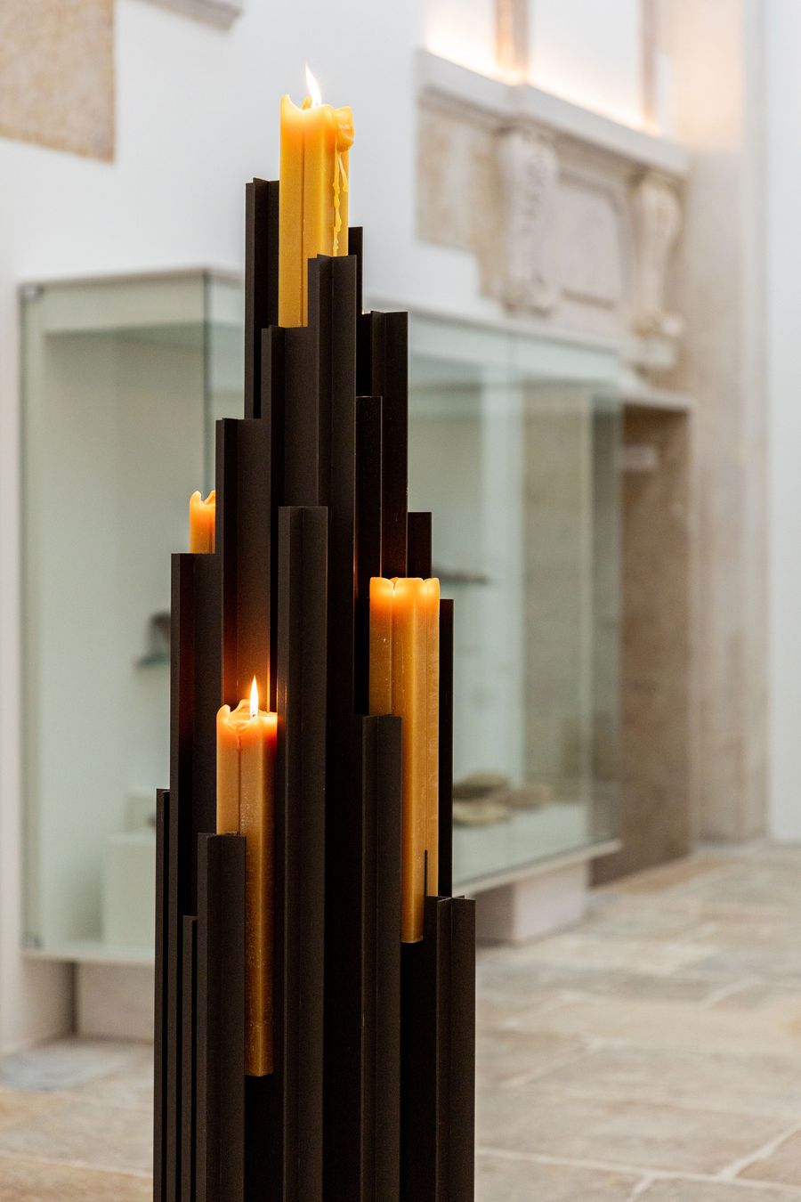 A dramatic close-up of a sculptural candle holder by AB and AC Architects from the "Alma Mater" exhibition at Locke Santa Joana. Tall, dark, vertical fins of varying heights hold glowing amber candles, creating an architectural play of light and shadow. The design evokes a sense of solemnity and contemporary brutalism within the historic setting. A dramatic close-up of a sculptural candle holder by AB and AC Architects from the "Alma Mater" exhibition at Locke Santa Joana. Tall, dark, vertical fins of varying heights hold glowing amber candles, creating an architectural play of light and shadow. The design evokes a sense of solemnity and contemporary brutalism within the historic setting.