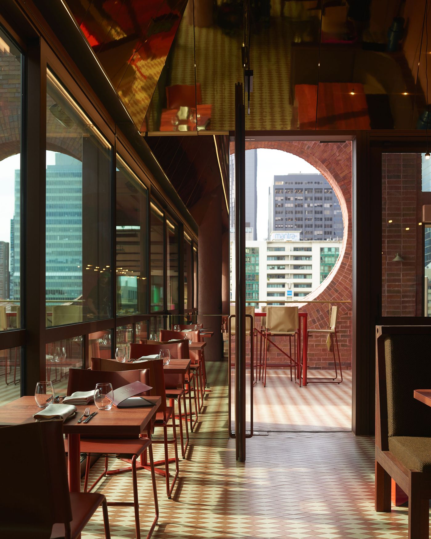 Modern restaurant interior with wooden tables, glass walls, and an open view of the city skyline through a circular window.
