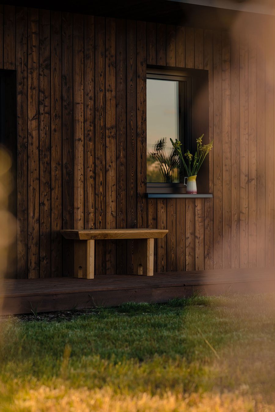 A wooden bench sits beside a modern wooden wall, with a window displaying a vase of flowers against a sunset backdrop. A wooden bench sits beside a modern wooden wall, with a window displaying a vase of flowers against a sunset backdrop.