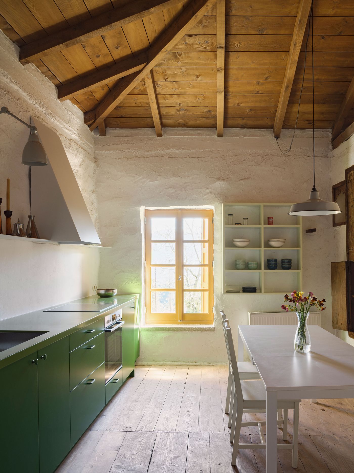 Exposed timber roof structure creates rhythmic patterns above the sage-green kitchen, where industrial pendant lighting and open shelving provide modern functionality. The composition balances traditional construction methods with contemporary design sensibilities, demonstrating how heritage buildings can accommodate modern domestic requirements through respectful intervention.