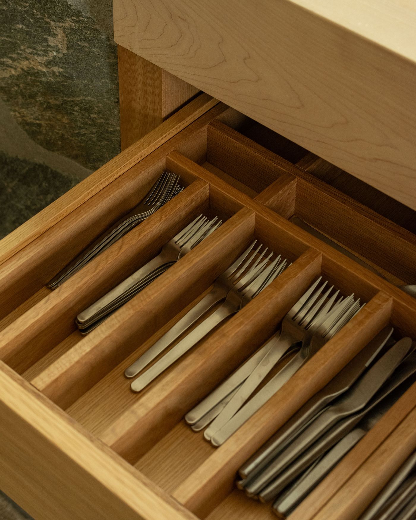 A wooden drawer open to reveal neatly organized cutlery, including forks and knives, in a clean, modern kitchen setting.