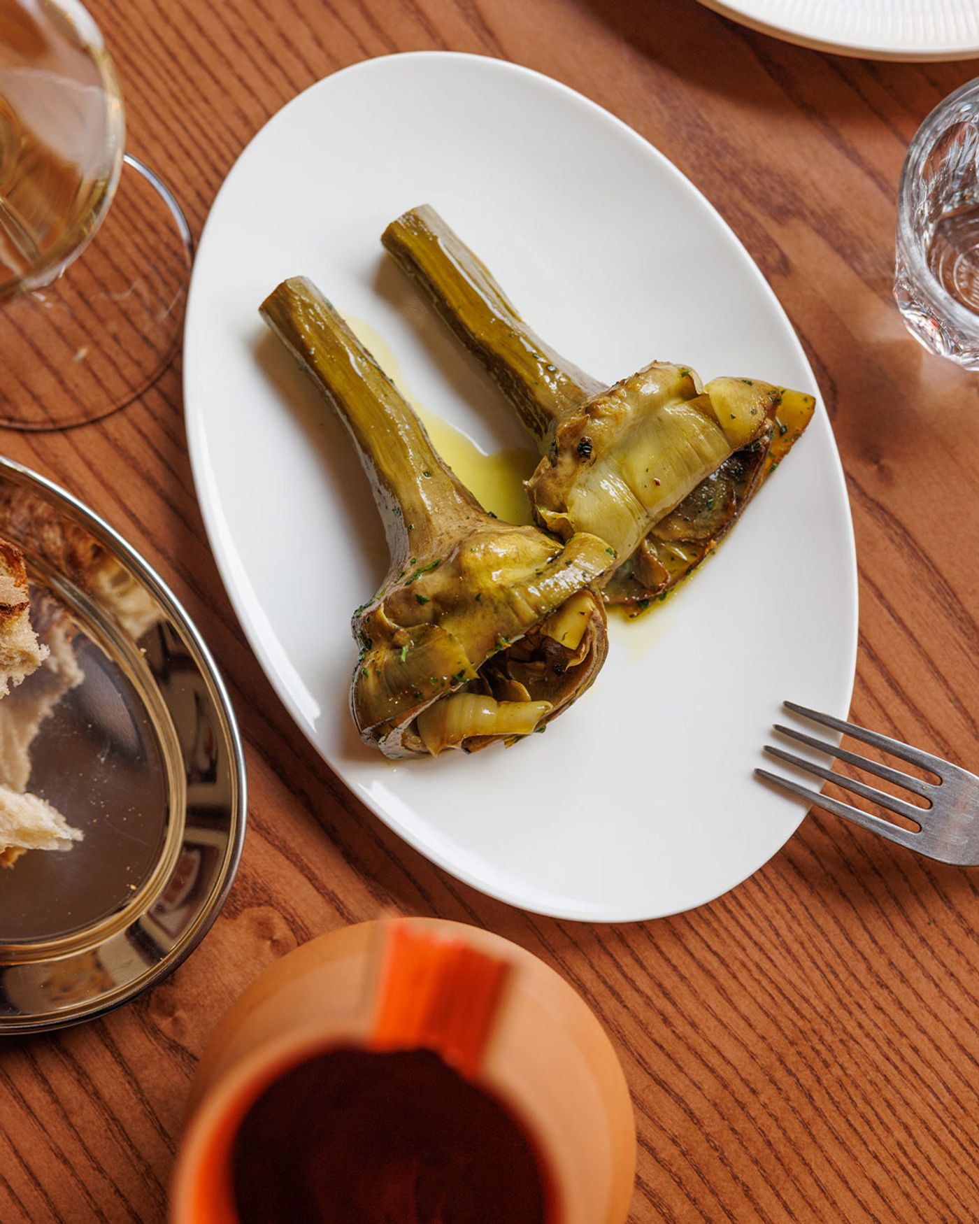 A close-up of a rustic yet refined dish from Santo Palato: two tender artichokes, glistening with oil, presented on a white oval plate. A fork rests beside the plate, and a hint of bread and a glass of wine are visible, suggesting a delightful and authentic Roman culinary experience.