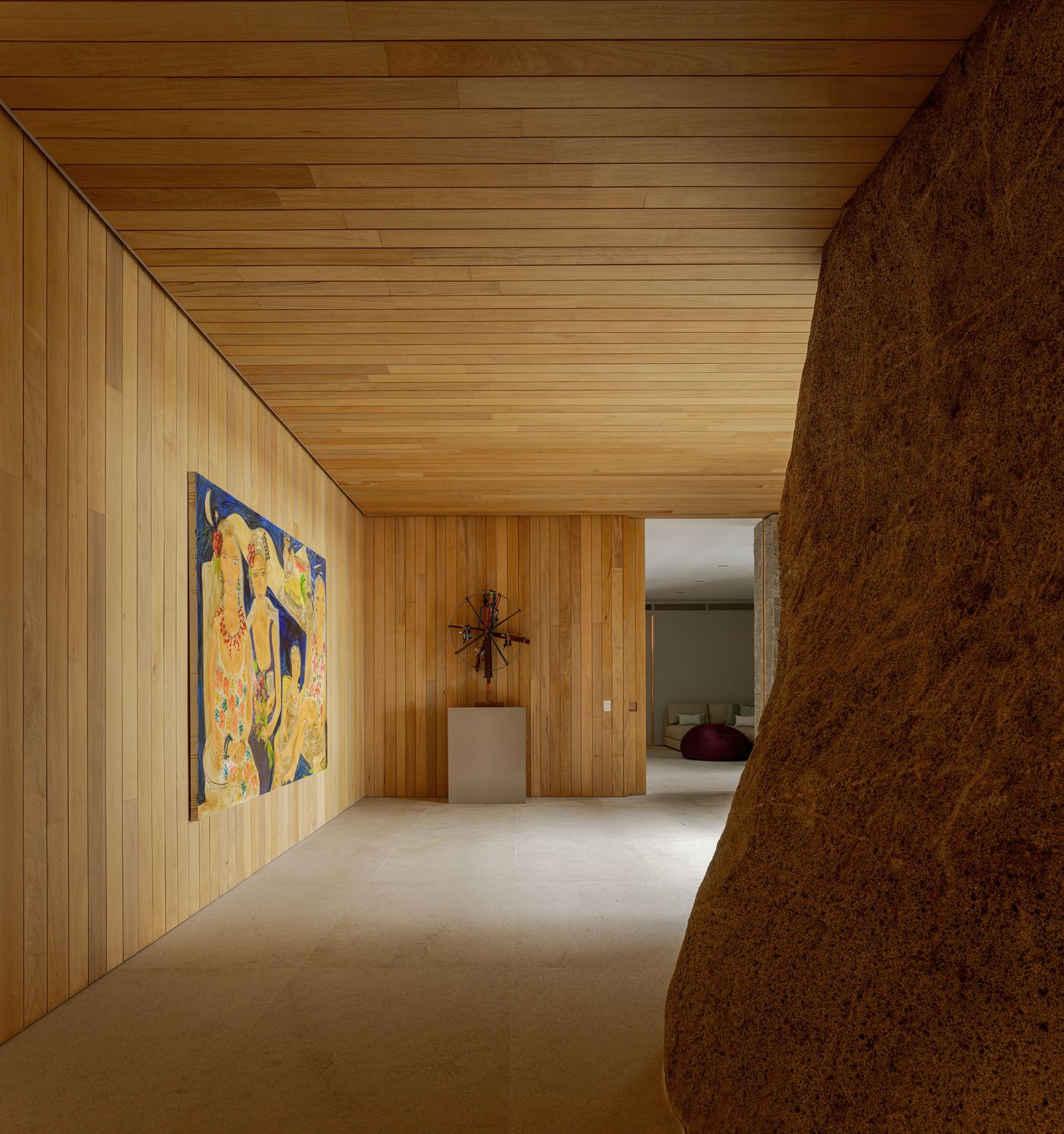 A minimalist interior hallway features warm wooden paneling on walls and ceiling, providing a serene backdrop for vibrant contemporary art. A massive, raw rock formation dramatically intersects the space, showcasing the dwelling's commitment to integrating natural elements and creating a unique, authentic atmosphere.