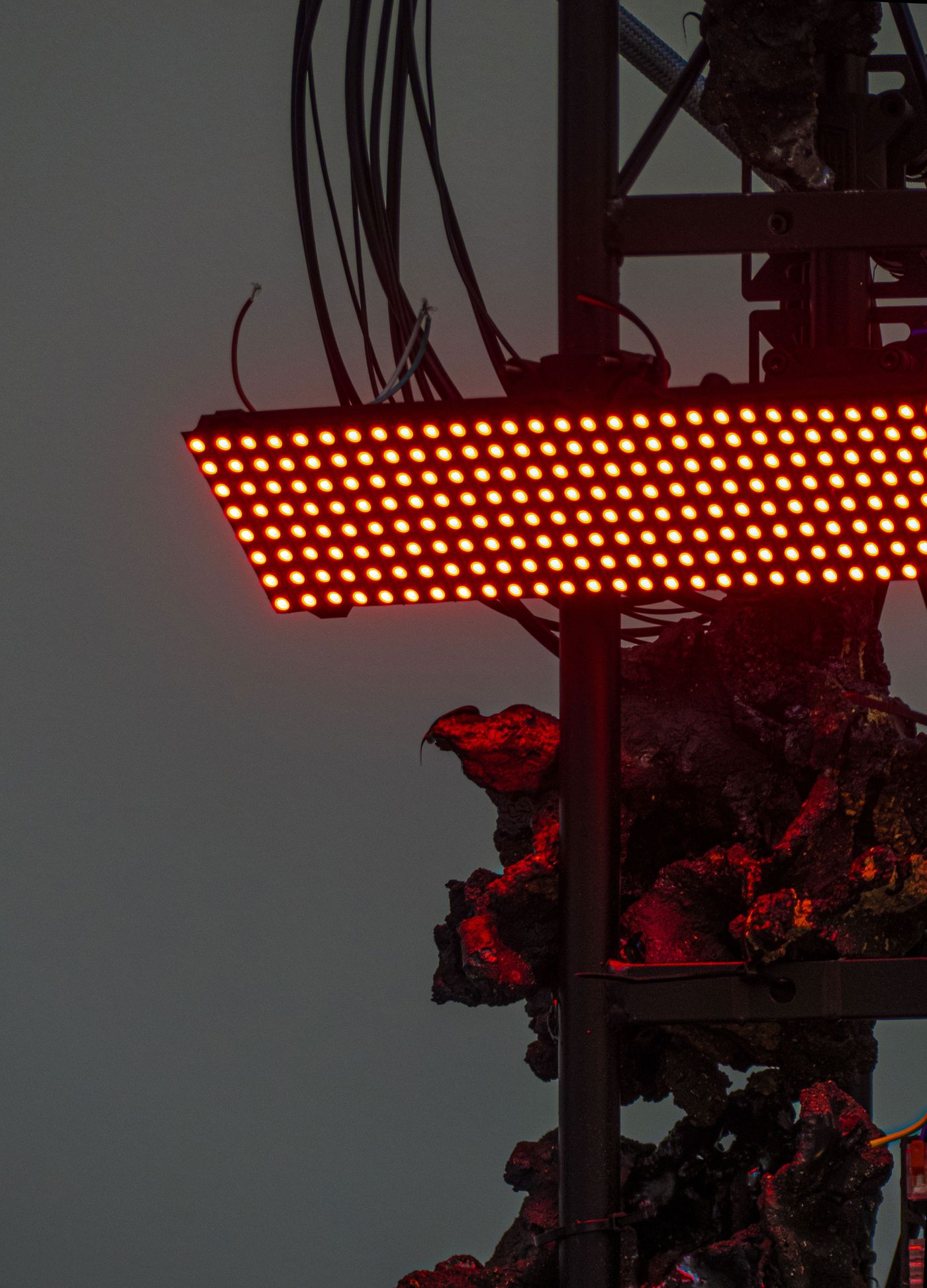 A close-up of a red LED display board mounted on a dark, textured structure, with visible wires and cables above.