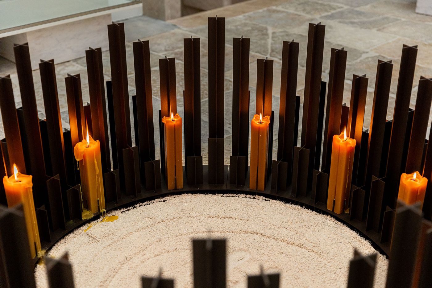 A close-up of the circular, ground-level candle installation from AB and AC Architects' "Alma Mater" exhibition at Locke Santa Joana. A ring of dark, vertical fin-like structures encircle a bed of light-colored sand, with warm amber candles flickering intermittently, creating a focal point of contemplative light and shadow against the textured stone floor. A close-up of the circular, ground-level candle installation from AB and AC Architects' "Alma Mater" exhibition at Locke Santa Joana. A ring of dark, vertical fin-like structures encircle a bed of light-colored sand, with warm amber candles flickering intermittently, creating a focal point of contemplative light and shadow against the textured stone floor.