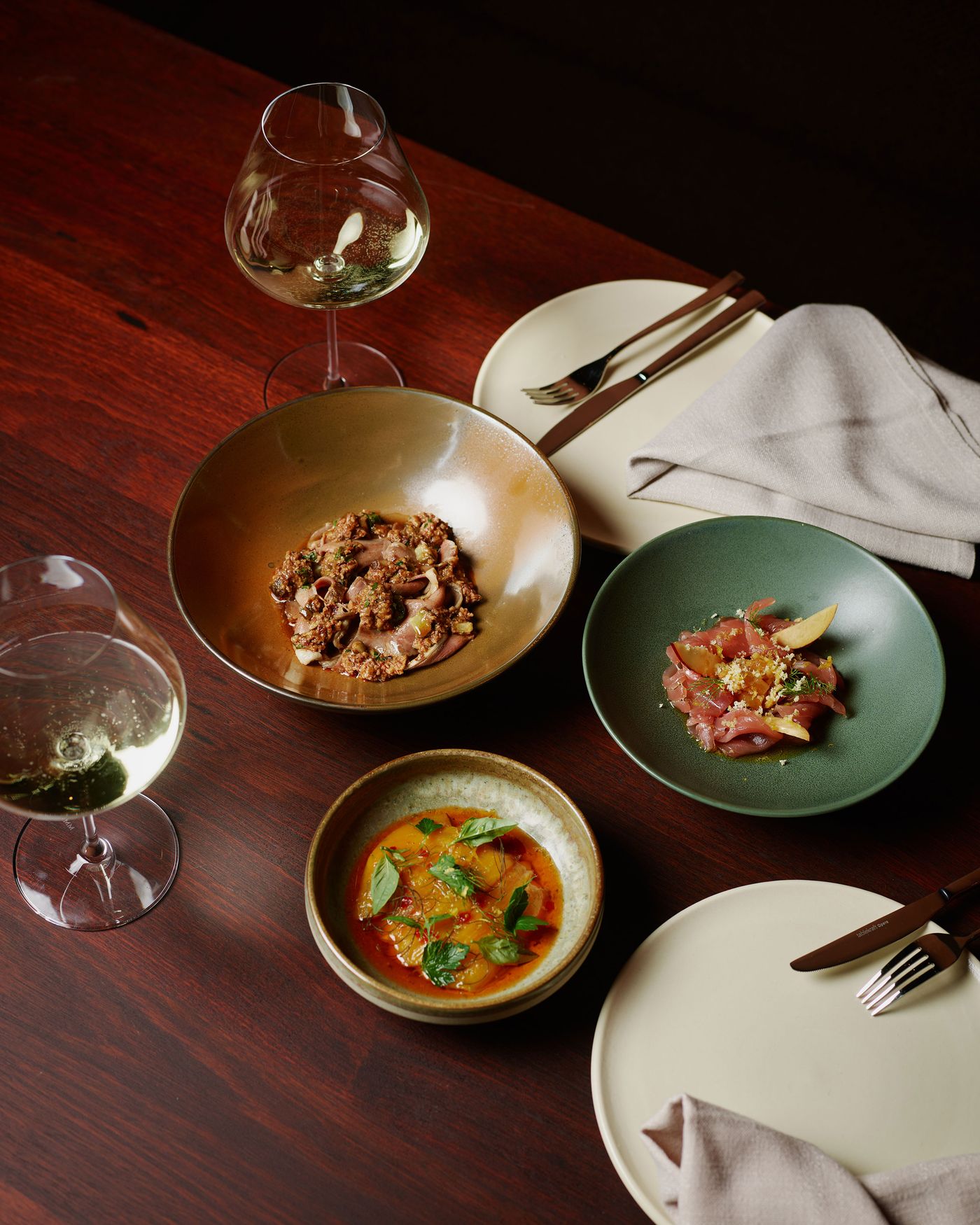 An elegant dining scene featuring a golden bowl of meat dish, a green plate with carpaccio, and a round bowl of citrus salad, with white wine.