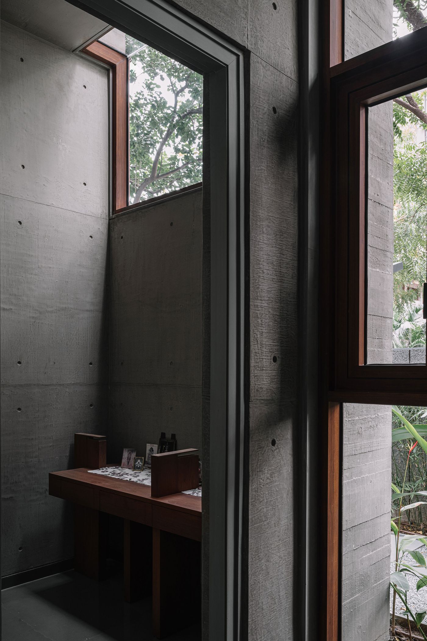 A quiet, contemplative nook, possibly a prayer room or study, with raw concrete walls textured by formwork. A simple wooden bench serves as furniture, bathed in natural light from a high window that frames a glimpse of tree branches. The minimalist design emphasizes the interplay of light and shadow on the robust materials.
