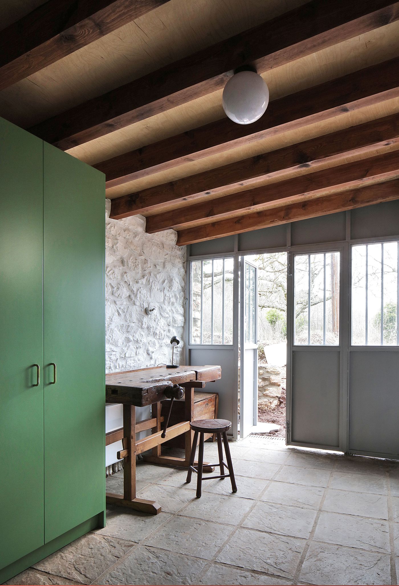 Traditional workbench becomes functional furniture within the sage-green contemporary framework, where expansive glazing connects interior workspace to Mediterranean landscape. The authentic craftsman's tools create material dialogue with modern cabinetry, celebrating both historical function and contemporary comfort within the restored stone structure.