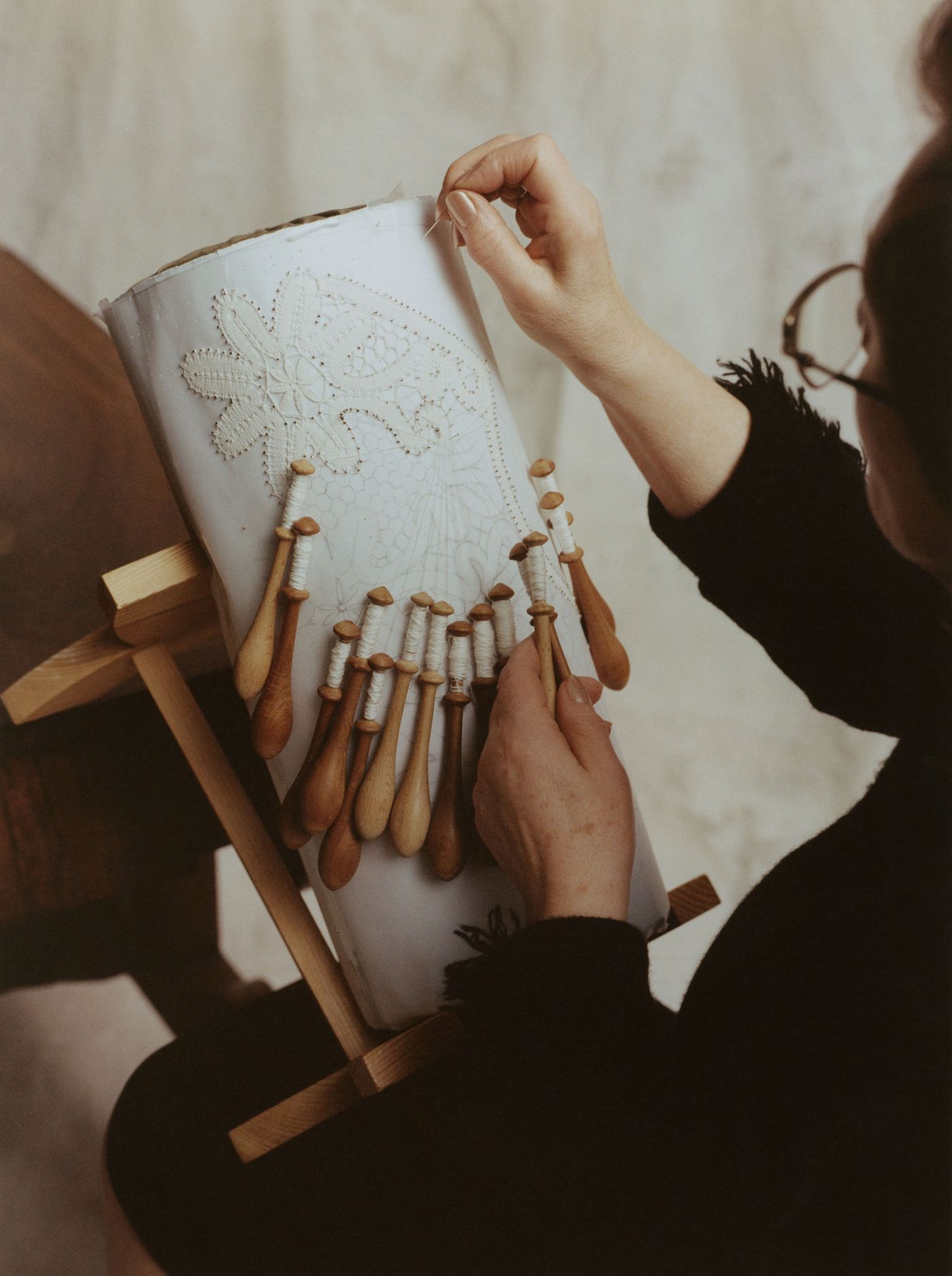 A close-up of a woman lace-making, threading a needle into intricate patterns on a white display, surrounded by wooden bobbins.