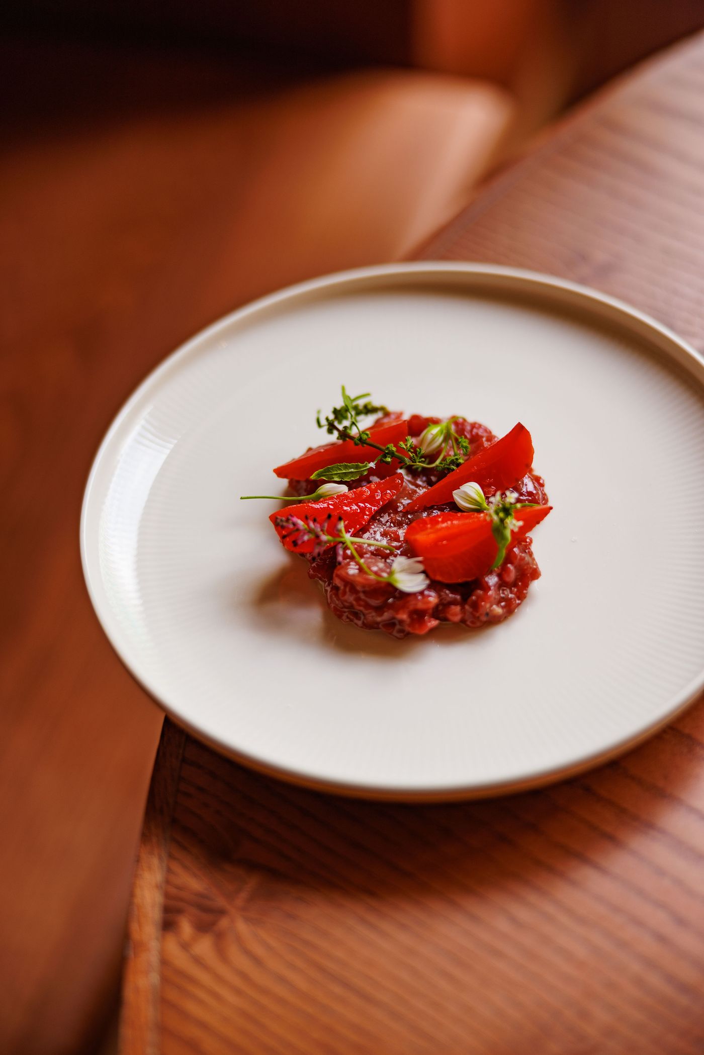 A vibrantly presented dish of beef tartare from Santo Palato, artfully arranged on a white, textured plate. The rich red meat is adorned with delicate green herbs, small white flowers, and slices of bright red tomato, showcasing a meticulous attention to detail and fresh ingredients.