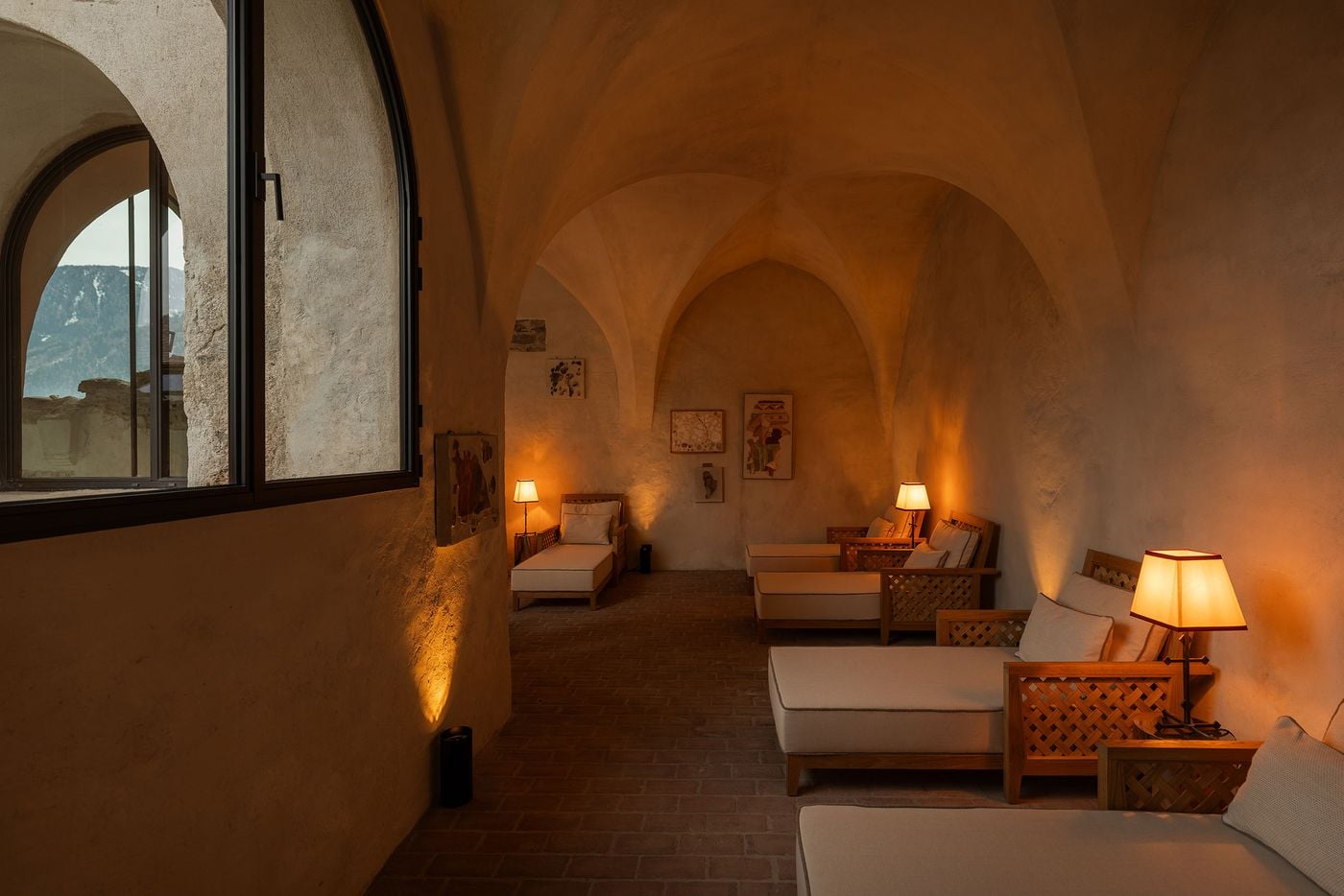 A spa relaxation room at Castel Badia occupies a softly lit, groin-vaulted chamber with a terracotta brick floor. Lattice-framed teak loungers with cream cushions line the walls beneath pleated lamps and framed works resembling fresco fragments, while an arched window looks out to medieval ruins and wooded slopes. A spa relaxation room at Castel Badia occupies a softly lit, groin-vaulted chamber with a terracotta brick floor. Lattice-framed teak loungers with cream cushions line the walls beneath pleated lamps and framed works resembling fresco fragments, while an arched window looks out to medieval ruins and wooded slopes.
