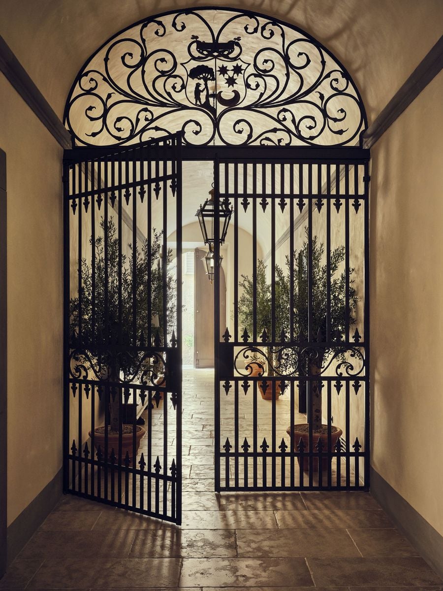 A wrought-iron gate at La Réserve Firenze, crowned by a scrolling lunette with a heraldic shield of a tree, stars and a crescent, stands half open onto the entrance passage. Beyond the bars, olive trees in terracotta pots and hanging lanterns lead the eye towards daylight from the street. A wrought-iron gate at La Réserve Firenze, crowned by a scrolling lunette with a heraldic shield of a tree, stars and a crescent, stands half open onto the entrance passage. Beyond the bars, olive trees in terracotta pots and hanging lanterns lead the eye towards daylight from the street.