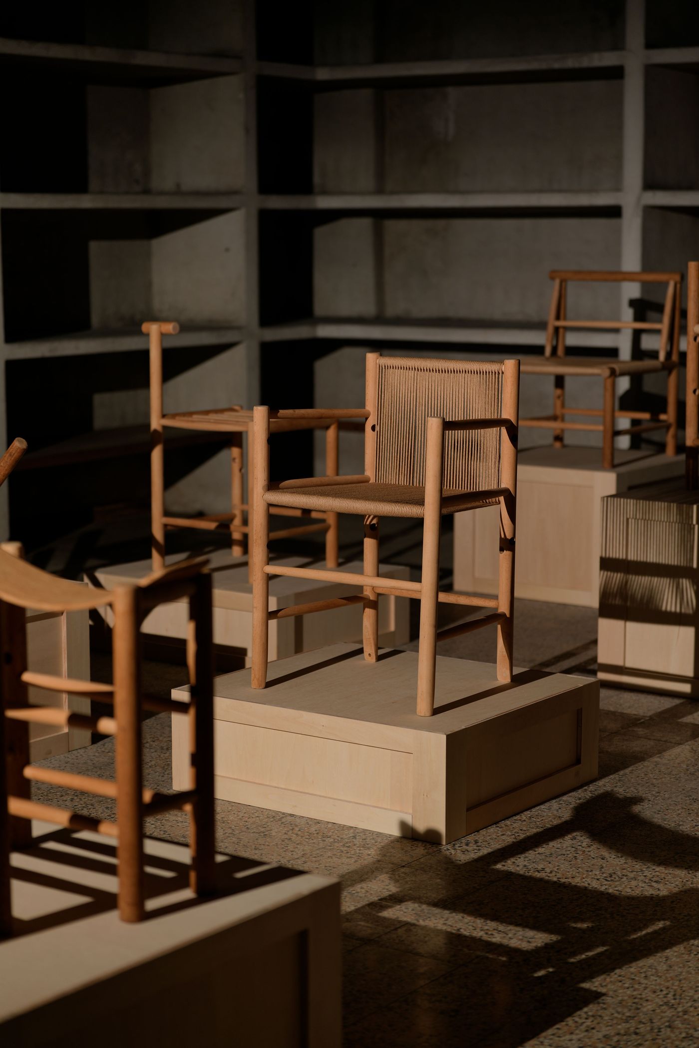 A collection of elegantly designed wooden chairs is displayed on light-colored pedestals in a dimly lit, modern setting. A collection of elegantly designed wooden chairs is displayed on light-colored pedestals in a dimly lit, modern setting.