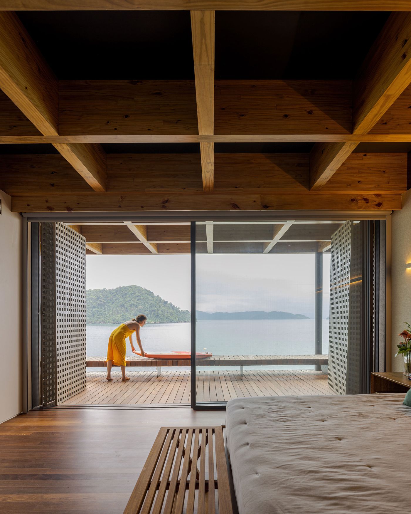A sun-drenched bedroom opens entirely to a private wooden deck, with a person in yellow looking out at the serene ocean and distant islands. The exposed timber ceiling and perforated sliding screens create a warm, inviting atmosphere, blending natural materials with a minimalist approach to maximize the stunning coastal views.