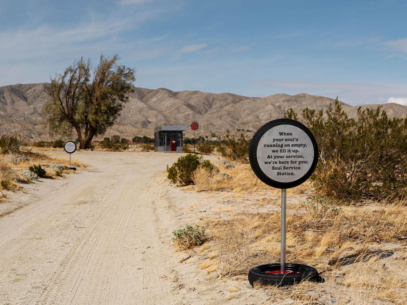A sign on a dirt road reads, "Soul Service Station," offering support with a desert backdrop and mountains in the distance. A sign on a dirt road reads, "Soul Service Station," offering support with a desert backdrop and mountains in the distance.