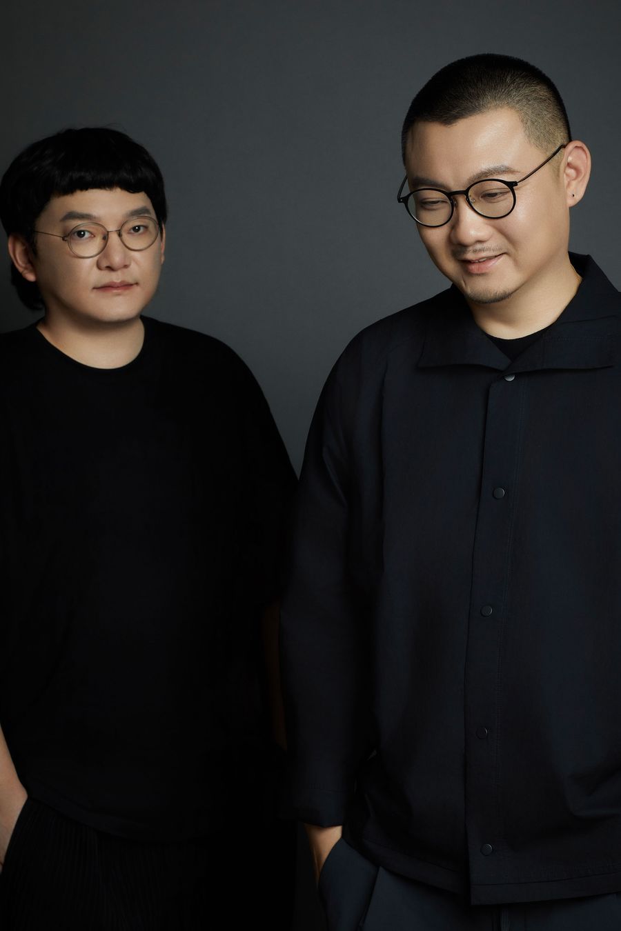 Two individuals dressed in black attire stand side by side against a dark background, exuding a modern, minimalist aesthetic. Two individuals dressed in black attire stand side by side against a dark background, exuding a modern, minimalist aesthetic.