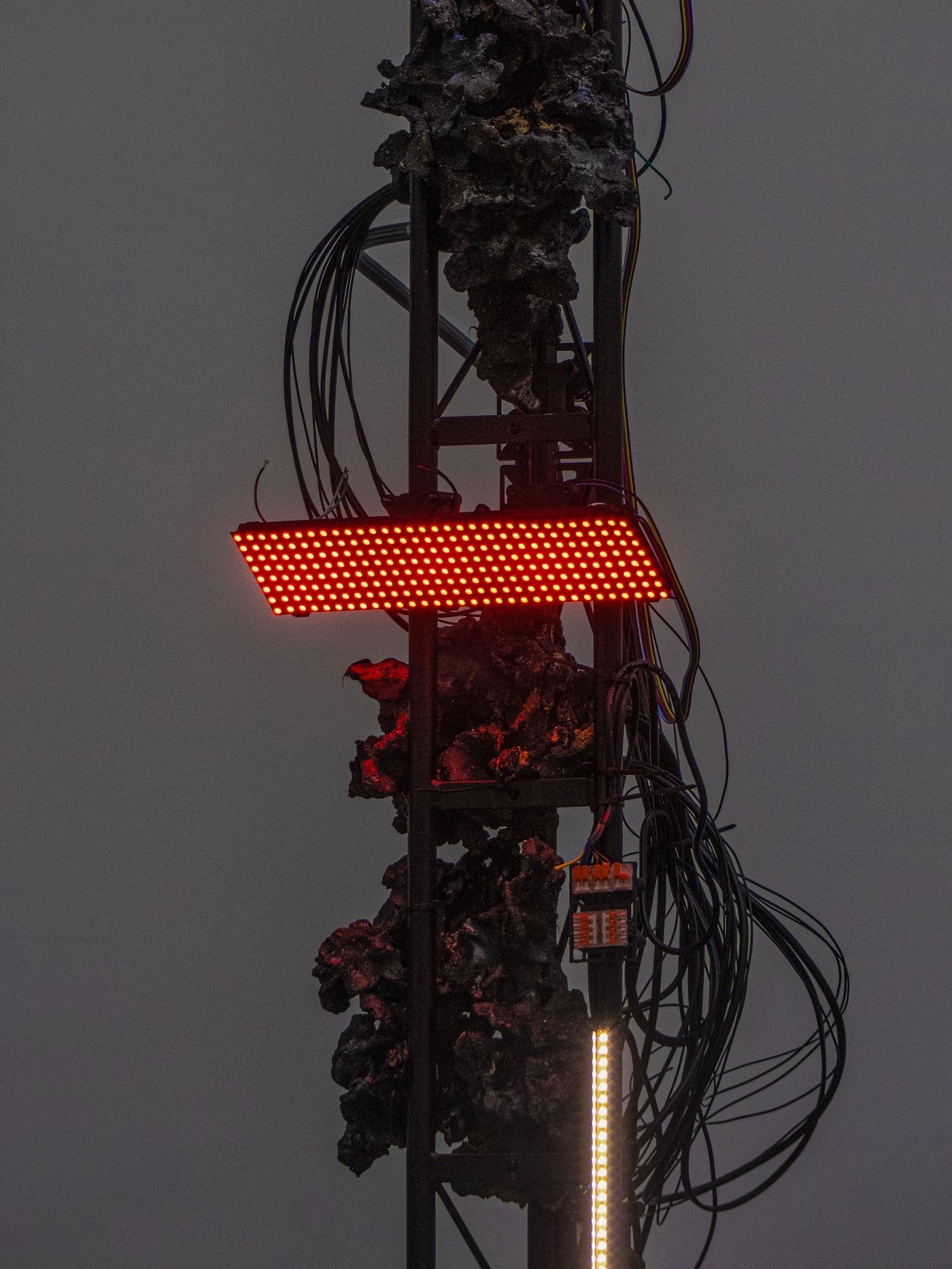 A metallic structure with a red light display, surrounded by tangled wires and a textured, dark surface resembling rock.