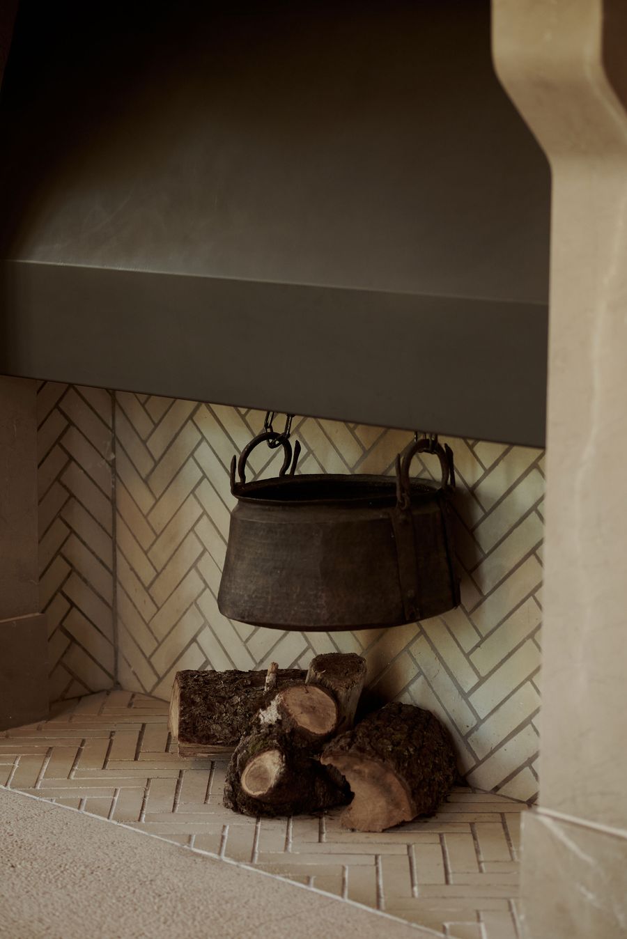 A close-up of a rustic-chic fireplace features a dark, antique-style cauldron suspended over a stack of natural firewood. The interior of the fireplace is lined with white, horizontally laid herringbone tiles, while the simple, elegant stone mantlepiece completes this inviting and authentic design element. A close-up of a rustic-chic fireplace features a dark, antique-style cauldron suspended over a stack of natural firewood. The interior of the fireplace is lined with white, horizontally laid herringbone tiles, while the simple, elegant stone mantlepiece completes this inviting and authentic design element.
