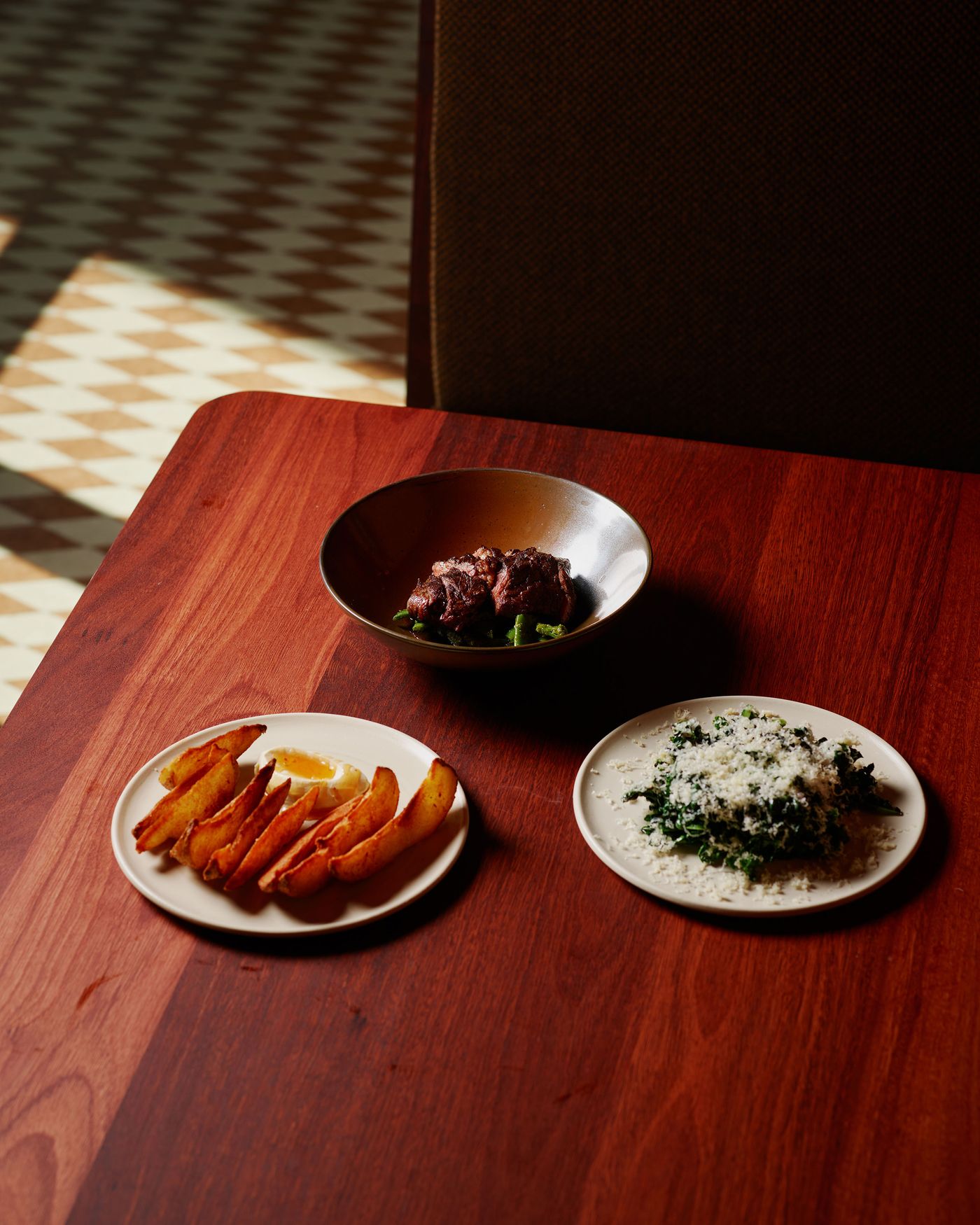 A wooden table features three dishes: tender meat with greens, crispy potato wedges, and a cheesy green salad, all beautifully presented.