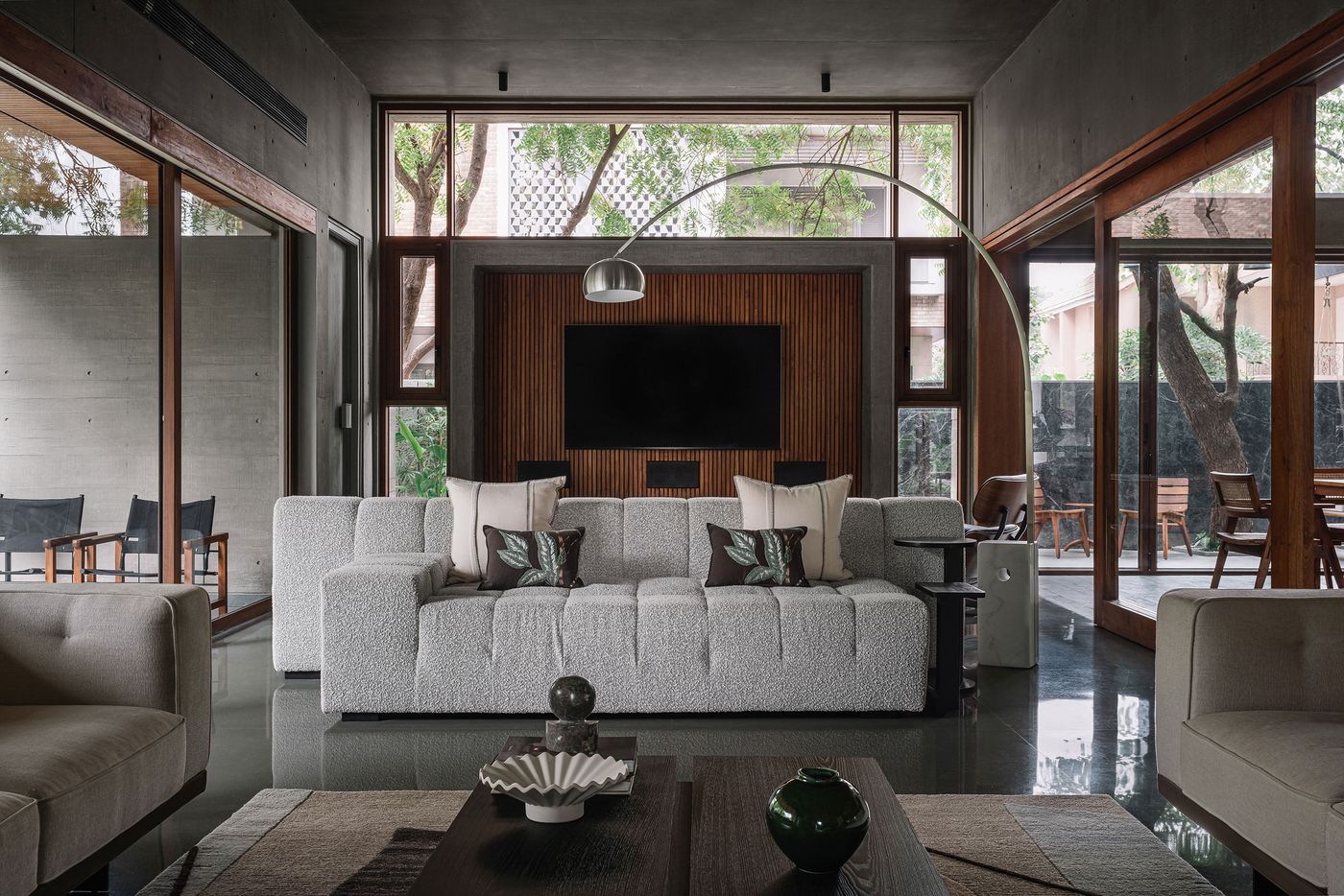 A sophisticated living room with two large, light grey modular sofas facing a prominent entertainment wall clad in warm wooden slats, featuring a large television. An arched floor lamp provides focused lighting. Expansive windows on either side offer views into lush courtyards, demonstrating a luxurious blend of minimalist design and biophilic connection.
