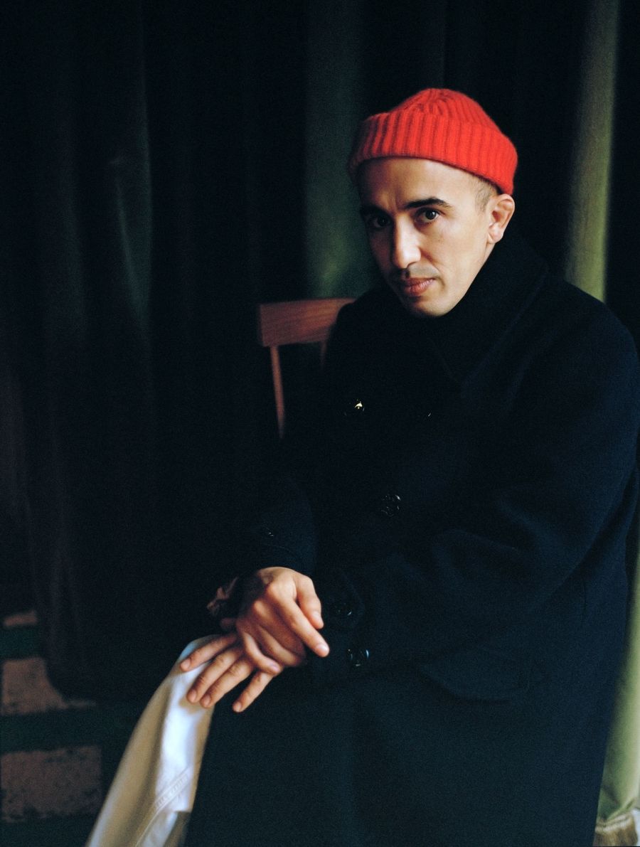 A person in a black coat and red beanie sits with hands clasped, surrounded by dark green curtains in a dimly lit setting.