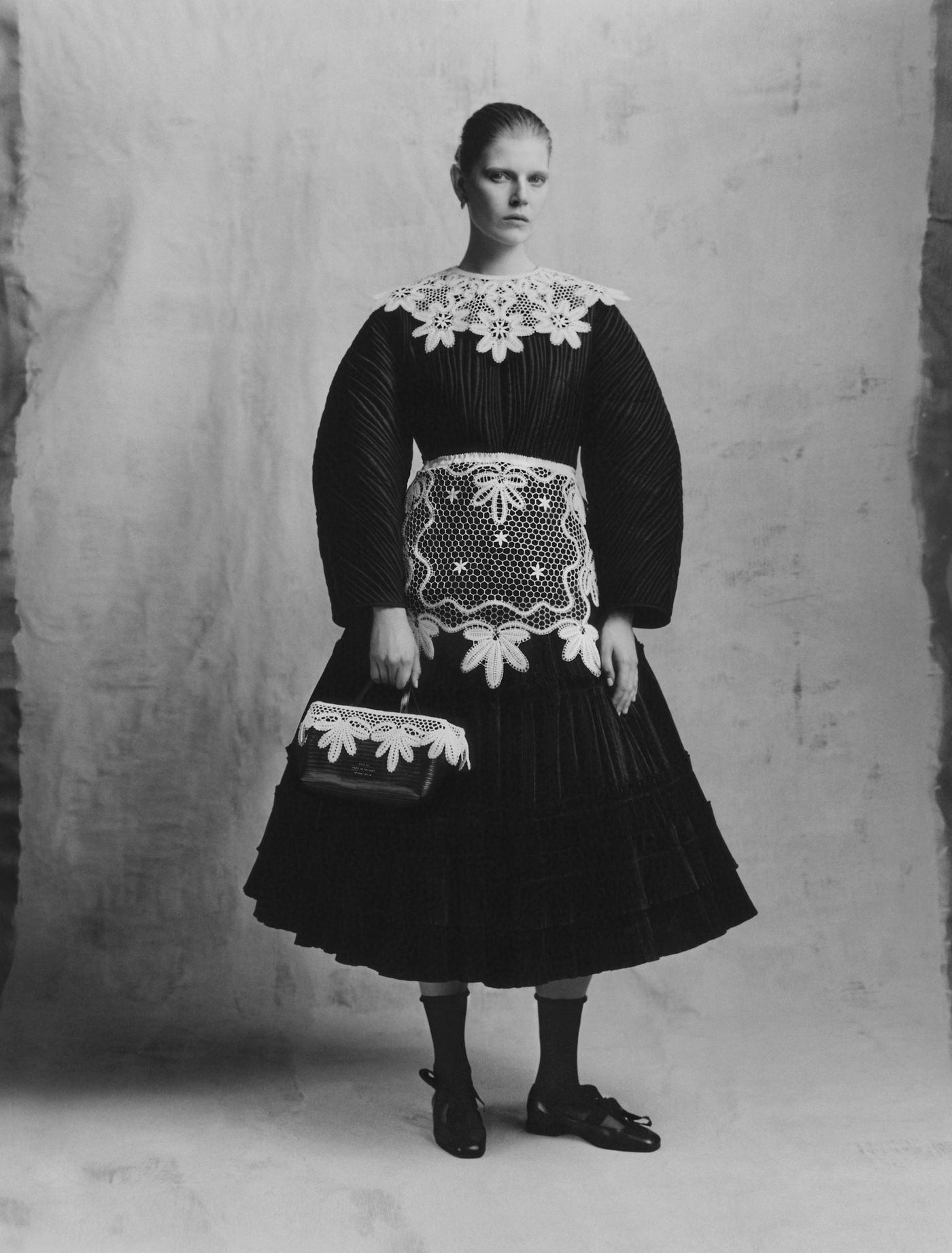 A figure in a traditional black dress with intricate white lace detailing and a matching handbag, standing against a neutral backdrop.