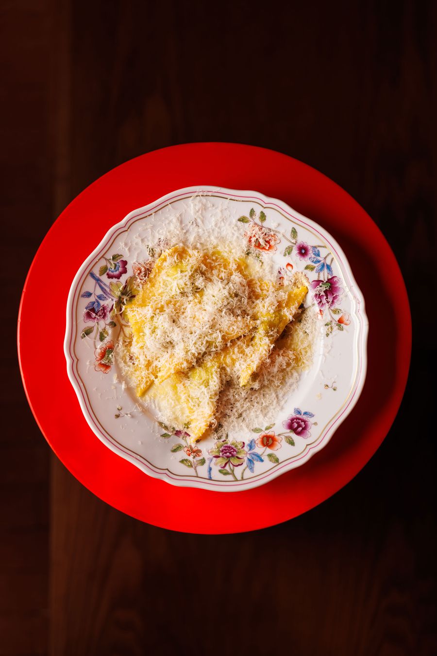 A visually rich plate of pasta, likely ravioli, from Santo Palato, generously covered in grated cheese and dark specks of pepper, served on a vintage-inspired floral porcelain plate. The plate rests on a bold, circular red charger, adding a vibrant contrast that emphasizes the dish's comforting and artful presentation. A visually rich plate of pasta, likely ravioli, from Santo Palato, generously covered in grated cheese and dark specks of pepper, served on a vintage-inspired floral porcelain plate. The plate rests on a bold, circular red charger, adding a vibrant contrast that emphasizes the dish's comforting and artful presentation.