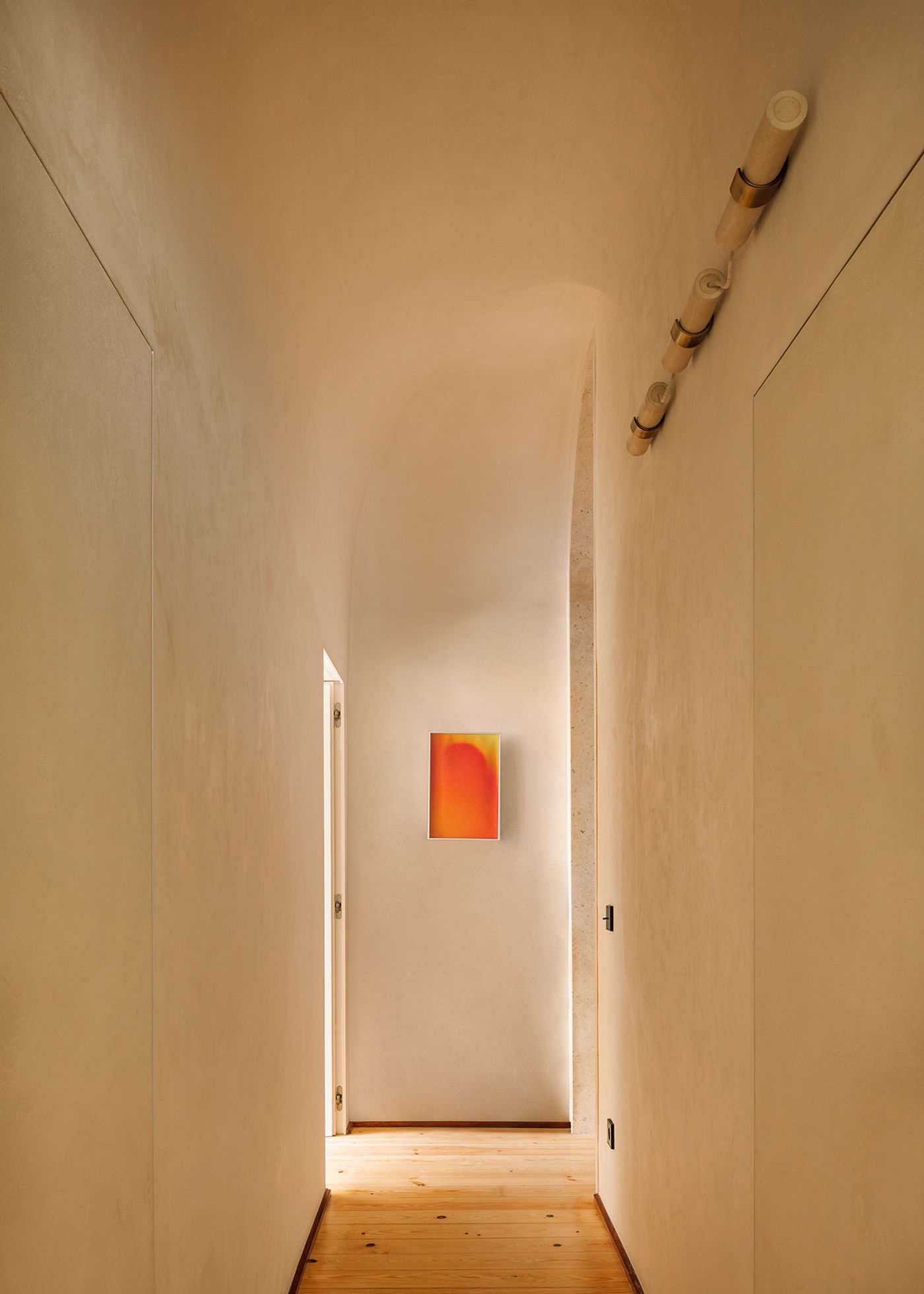 A softly lit hallway features smooth, beige walls, a wooden floor, and a vibrant orange artwork framed on the end wall. A softly lit hallway features smooth, beige walls, a wooden floor, and a vibrant orange artwork framed on the end wall.
