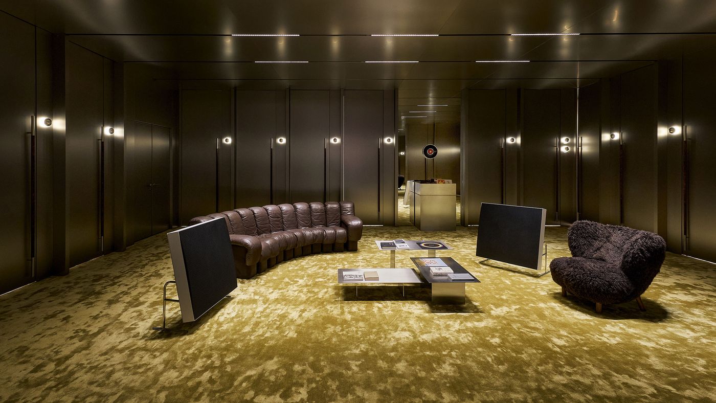 Modern lounge with curved brown leather sofa, a cozy dark chair, sleek tables, and warm lighting on textured gold carpet. Modern lounge with curved brown leather sofa, a cozy dark chair, sleek tables, and warm lighting on textured gold carpet.