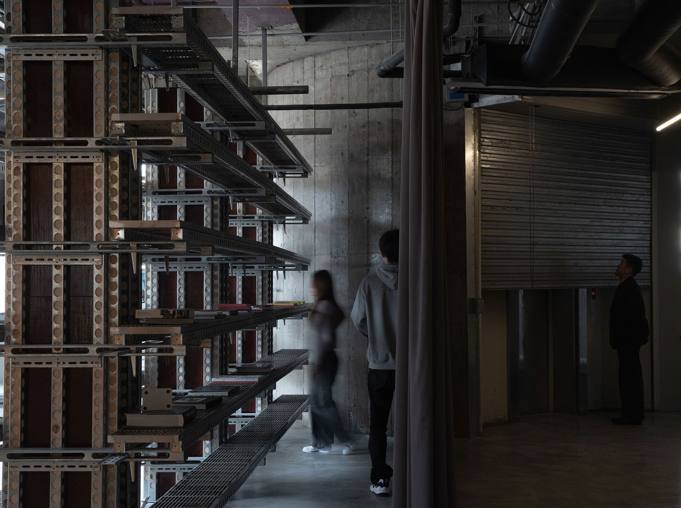 This image from 889 GLO Art Space focuses on the multi-layered industrial shelving system, showcasing its modular construction of metal and dark panels. Two blurred figures interact within the space, highlighting its scale. The raw concrete wall and a dark, corrugated metal surface in the background reinforce the robust, brutalist industrial aesthetic of the art space. This image from 889 GLO Art Space focuses on the multi-layered industrial shelving system, showcasing its modular construction of metal and dark panels. Two blurred figures interact within the space, highlighting its scale. The raw concrete wall and a dark, corrugated metal surface in the background reinforce the robust, brutalist industrial aesthetic of the art space.