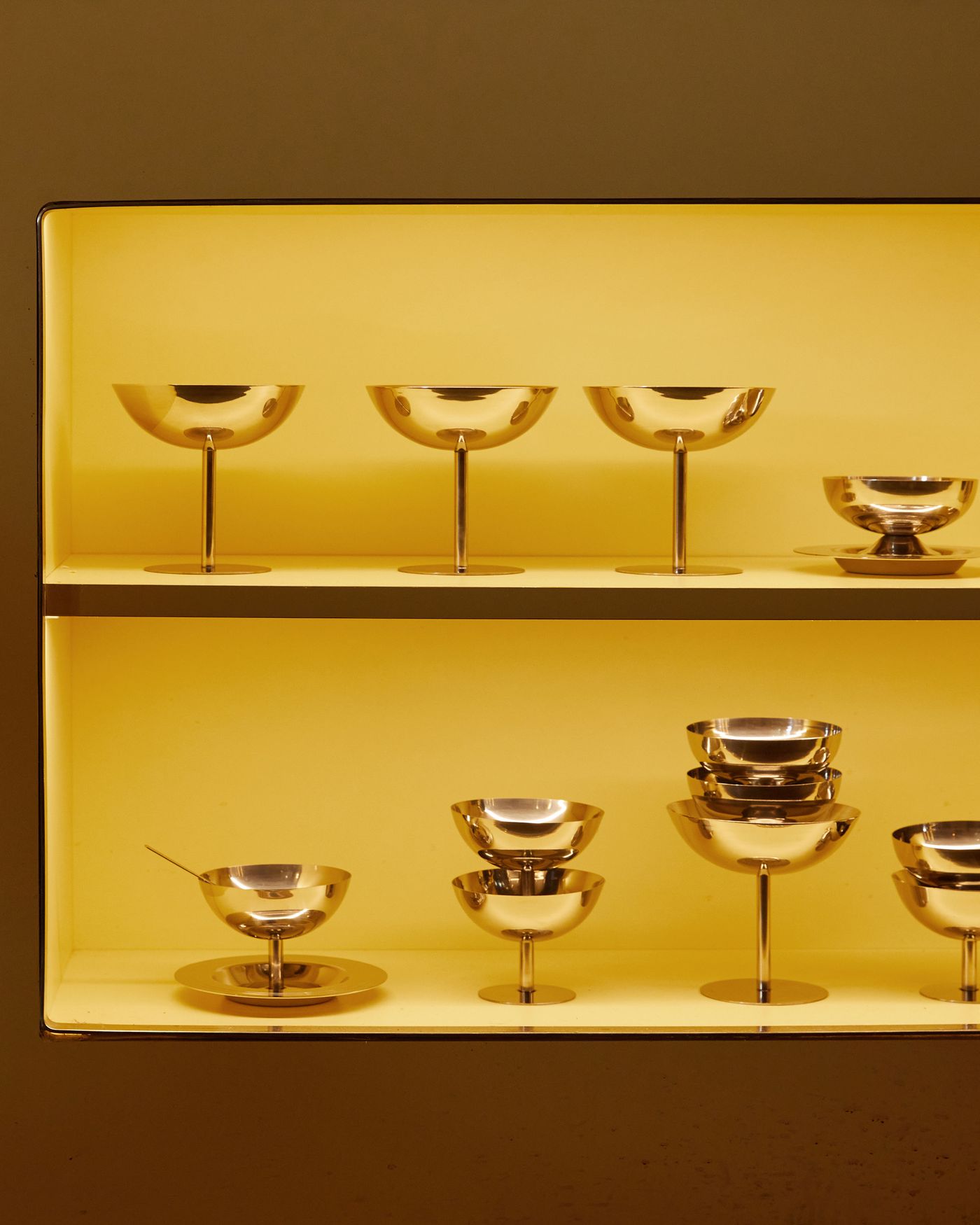 A vibrant culinary display features a shelving unit against a backdrop of glossy, yellow subway tiles. The warm light accentuates the metallic luster of various kitchen vessels and the clean, white ceramic tableware, creating a balanced composition of color, texture, and function.