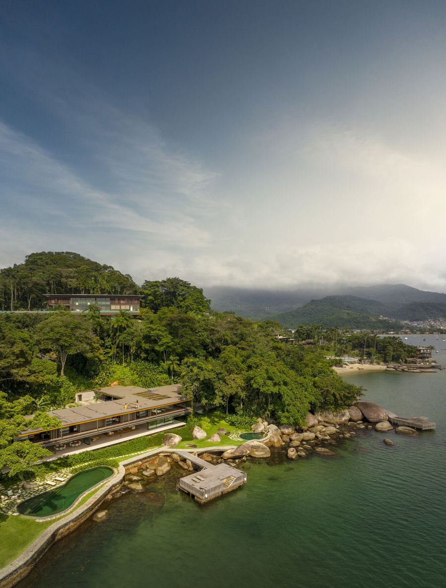An expansive aerial view captures a minimalist, horizontal dwelling on a lush, rocky peninsula, set amidst dense tropical foliage. The design incorporates natural pools and wooden decks that extend over the vibrant green-blue water, seamlessly connecting the residence with its stunning coastal environment. An expansive aerial view captures a minimalist, horizontal dwelling on a lush, rocky peninsula, set amidst dense tropical foliage. The design incorporates natural pools and wooden decks that extend over the vibrant green-blue water, seamlessly connecting the residence with its stunning coastal environment.