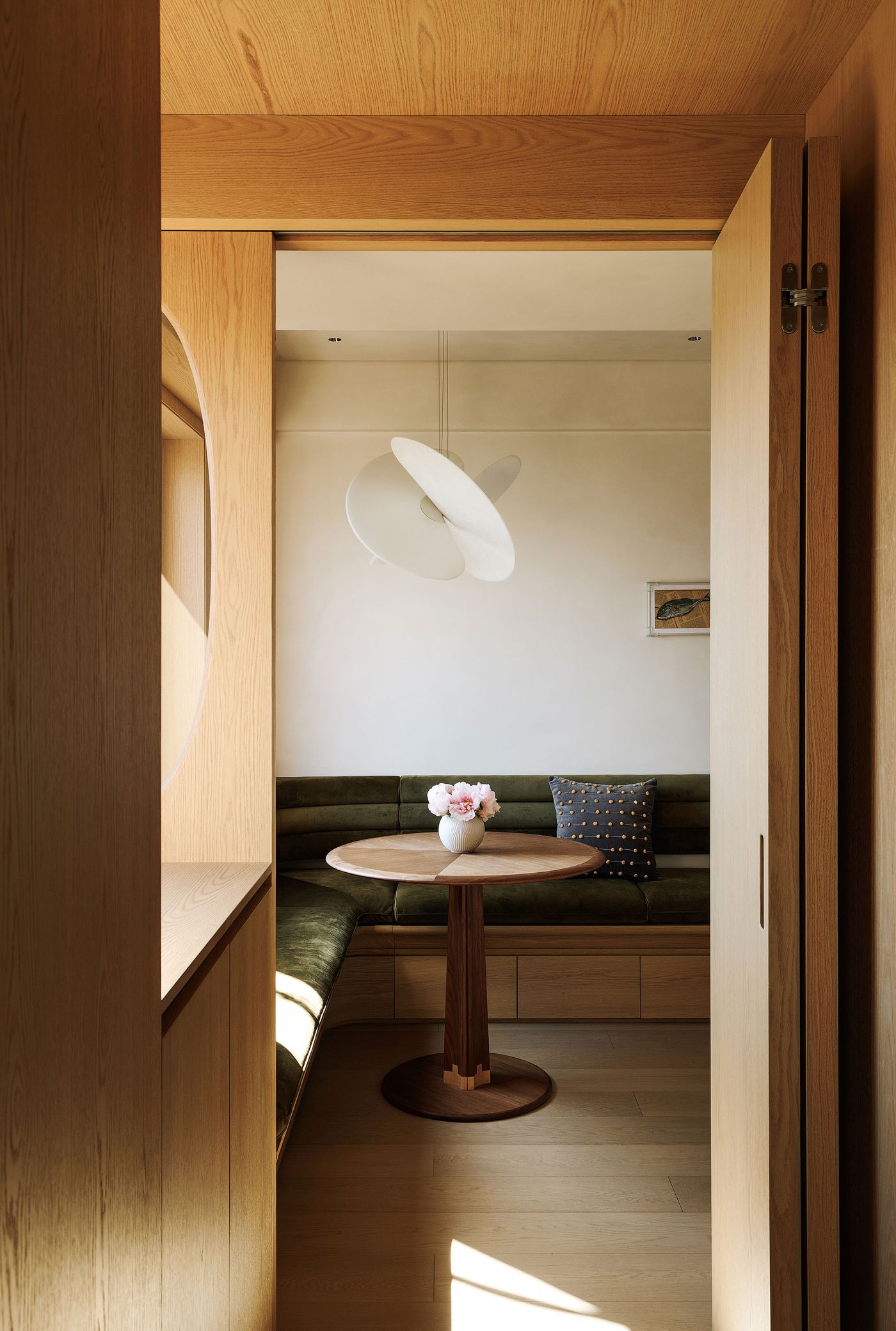 A cozy, Japandi-inspired dining nook with built-in olive green velvet seating nestled against warm wooden walls. A round wooden table with a unique base stands at the center, adorned with a delicate floral arrangement. The minimalist pendant light fixture above adds a sculptural element, enhancing the intimate and inviting atmosphere of this Shanghai apartment. A cozy, Japandi-inspired dining nook with built-in olive green velvet seating nestled against warm wooden walls. A round wooden table with a unique base stands at the center, adorned with a delicate floral arrangement. The minimalist pendant light fixture above adds a sculptural element, enhancing the intimate and inviting atmosphere of this Shanghai apartment.