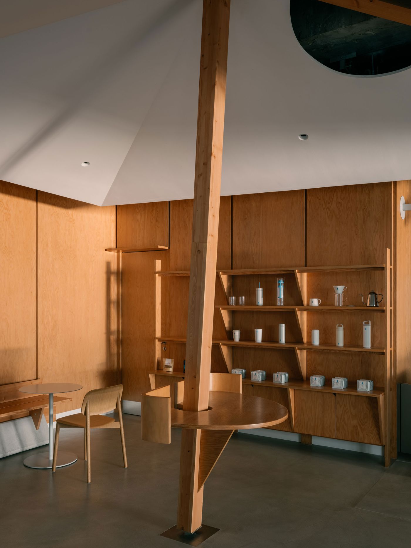 A quiet corner of the café reveals built-in oak shelving with carefully arranged ceramics and a unique circular table anchored to a central timber column, highlighting the space’s crafted simplicity.