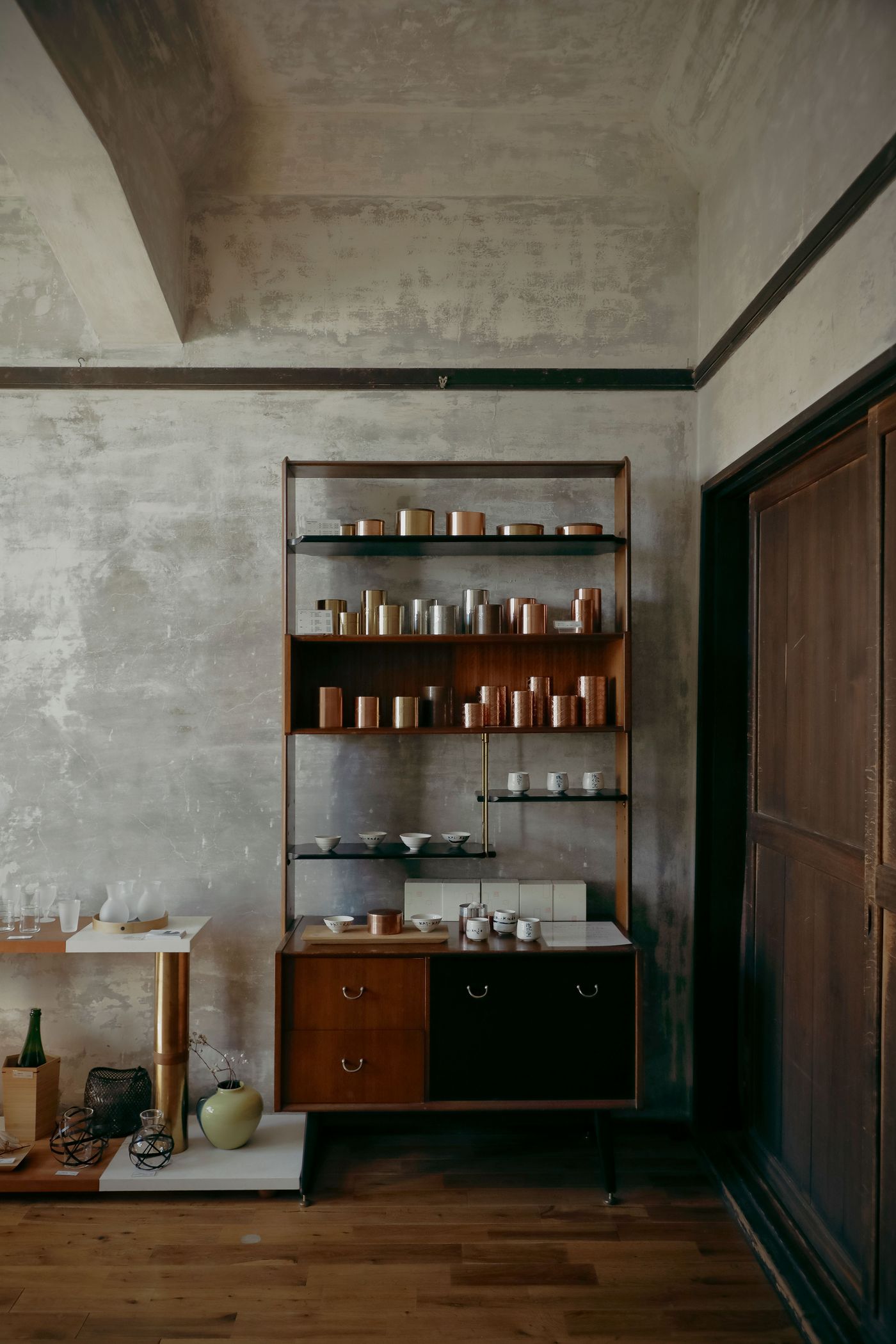 A wooden shelving unit display with various metallic containers and ceramic dishes, set against a textured gray wall and dark doorway. A wooden shelving unit display with various metallic containers and ceramic dishes, set against a textured gray wall and dark doorway.