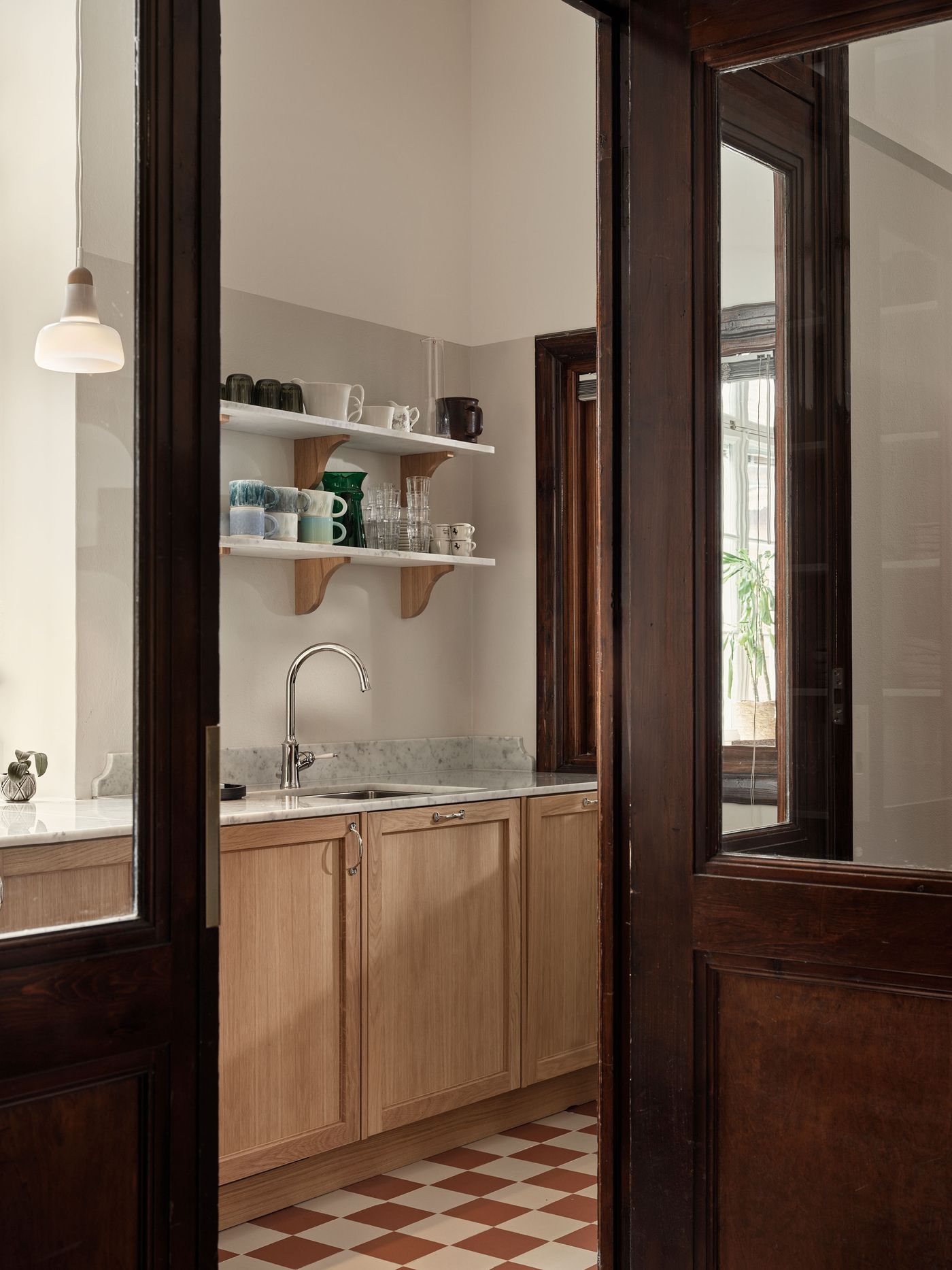 Through dark wooden framed glass doors, a contemporary kitchen space is revealed, featuring light wood cabinetry, a marble countertop with a sink, and open shelving. The red and white checkerboard floor extends into this functional area, maintaining the interior's distinctive pattern. Through dark wooden framed glass doors, a contemporary kitchen space is revealed, featuring light wood cabinetry, a marble countertop with a sink, and open shelving. The red and white checkerboard floor extends into this functional area, maintaining the interior's distinctive pattern.
