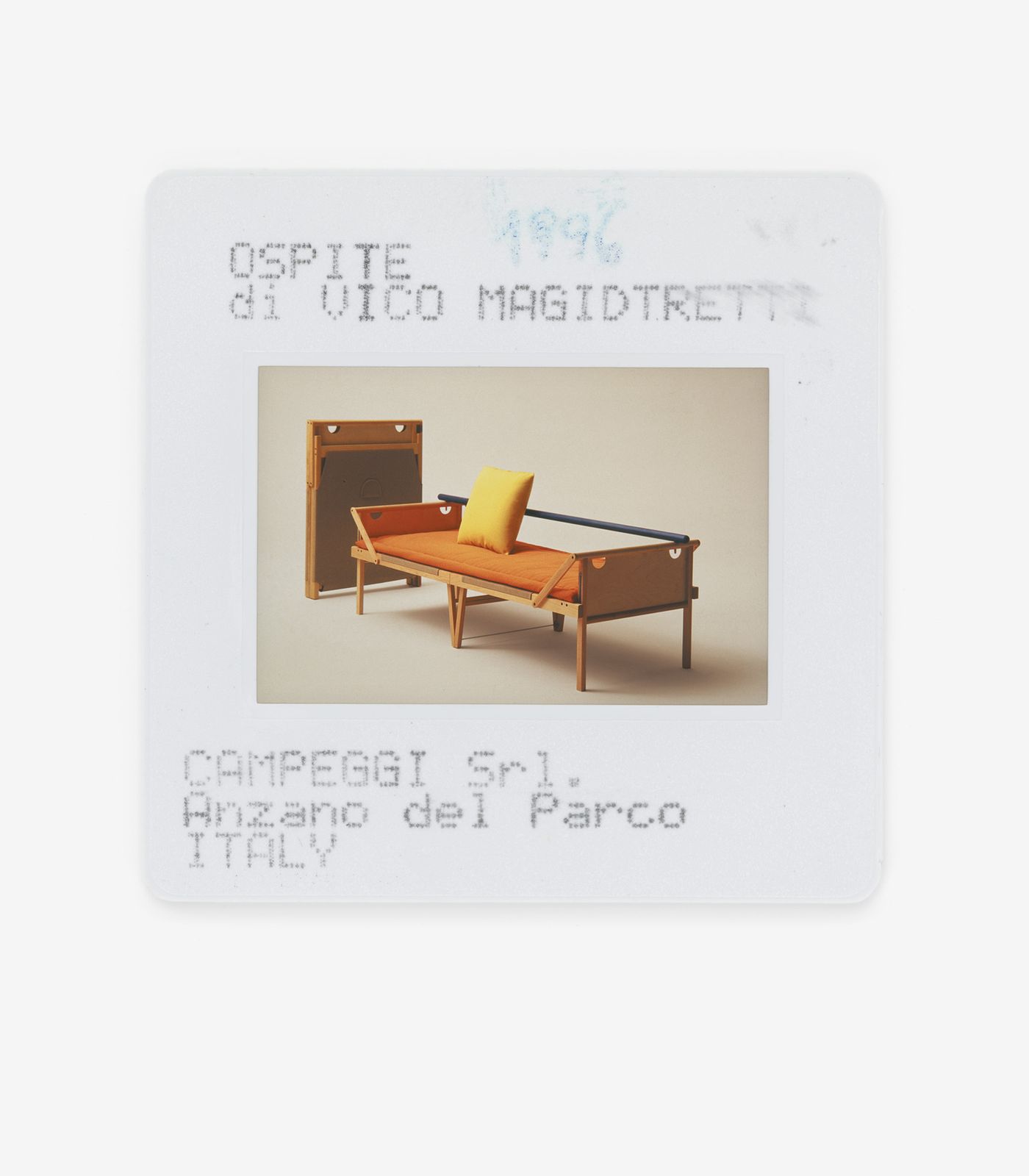 This image is a photograph of a slide, featuring a midcentury-modern daybed with an orange cushion and yellow accent pillow. The wooden frame of the bed is simple yet elegant, with clean lines. To the left, a disassembled or folded frame is visible. The text on the slide identifies the piece as "Ospite di Vico Magistretti" by CAMPEGGI Srl, Anzano del Parco, ITALY, dated 1996. The overall mood is retro and sophisticated.