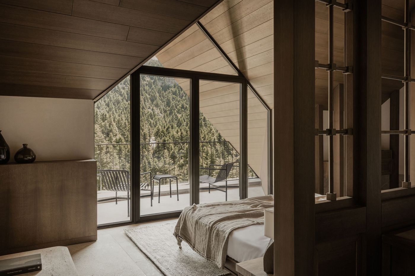 A cozy, yet expansive, bedroom embraces a contemporary rustic aesthetic with a vaulted timber ceiling and light wood paneling. Floor-to-ceiling windows frame a dramatic view of a dense, verdant forest, while a private balcony with minimalist chairs extends the living space outdoors, fostering a deep connection with nature.
