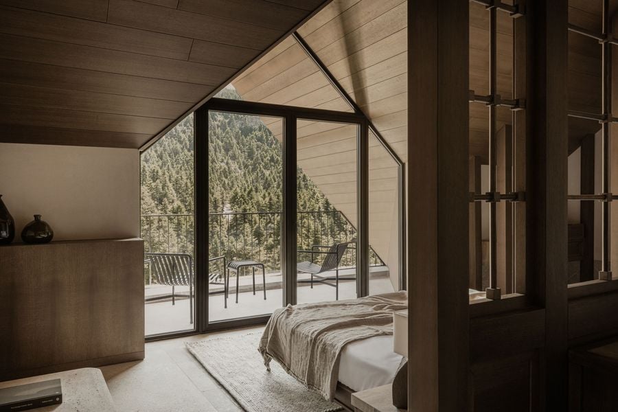 A cozy, yet expansive, bedroom embraces a contemporary rustic aesthetic with a vaulted timber ceiling and light wood paneling. Floor-to-ceiling windows frame a dramatic view of a dense, verdant forest, while a private balcony with minimalist chairs extends the living space outdoors, fostering a deep connection with nature.